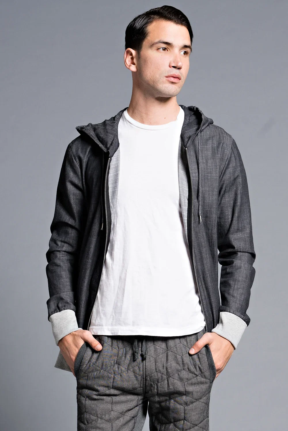 A young man with black hair wearing a white t-shirt, grey plaid pants, and a dark grey zip-up hoodie with puffy sleeves, standing against a plain grey background.
