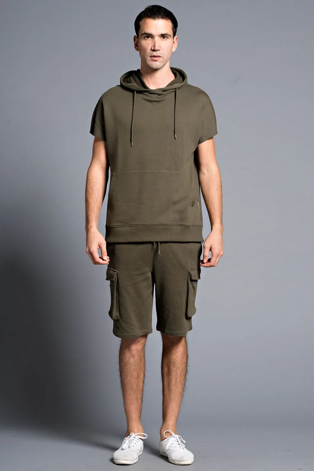 A young man with dark hair standing against a plain gray background, wearing an olive green hooded T-shirt, matching shorts with cargo pockets, and white sneakers.