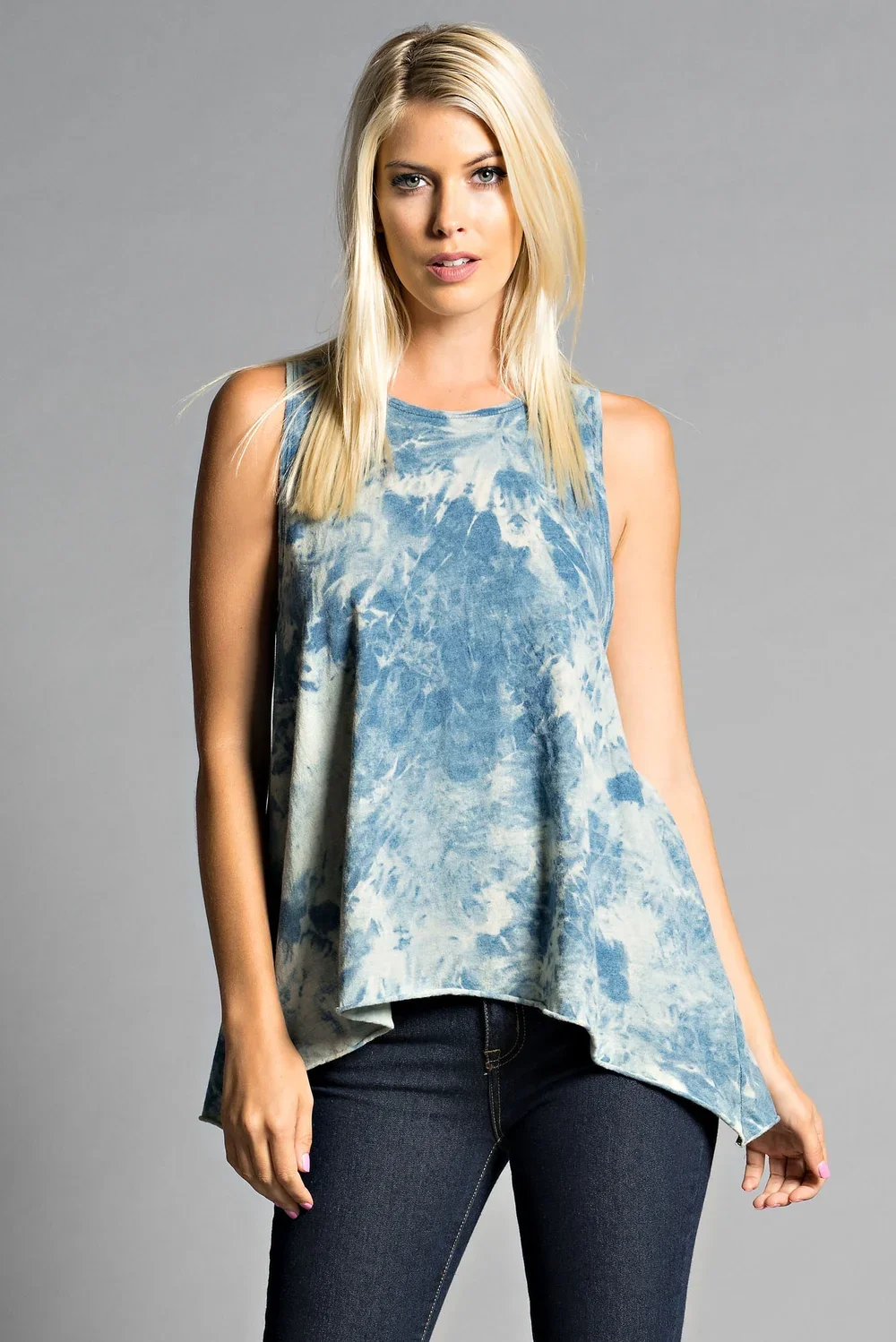A young woman with blonde hair and blue eyes wearing a sleeveless blue and white tie-dye top and dark jeans, standing against a plain gray background.