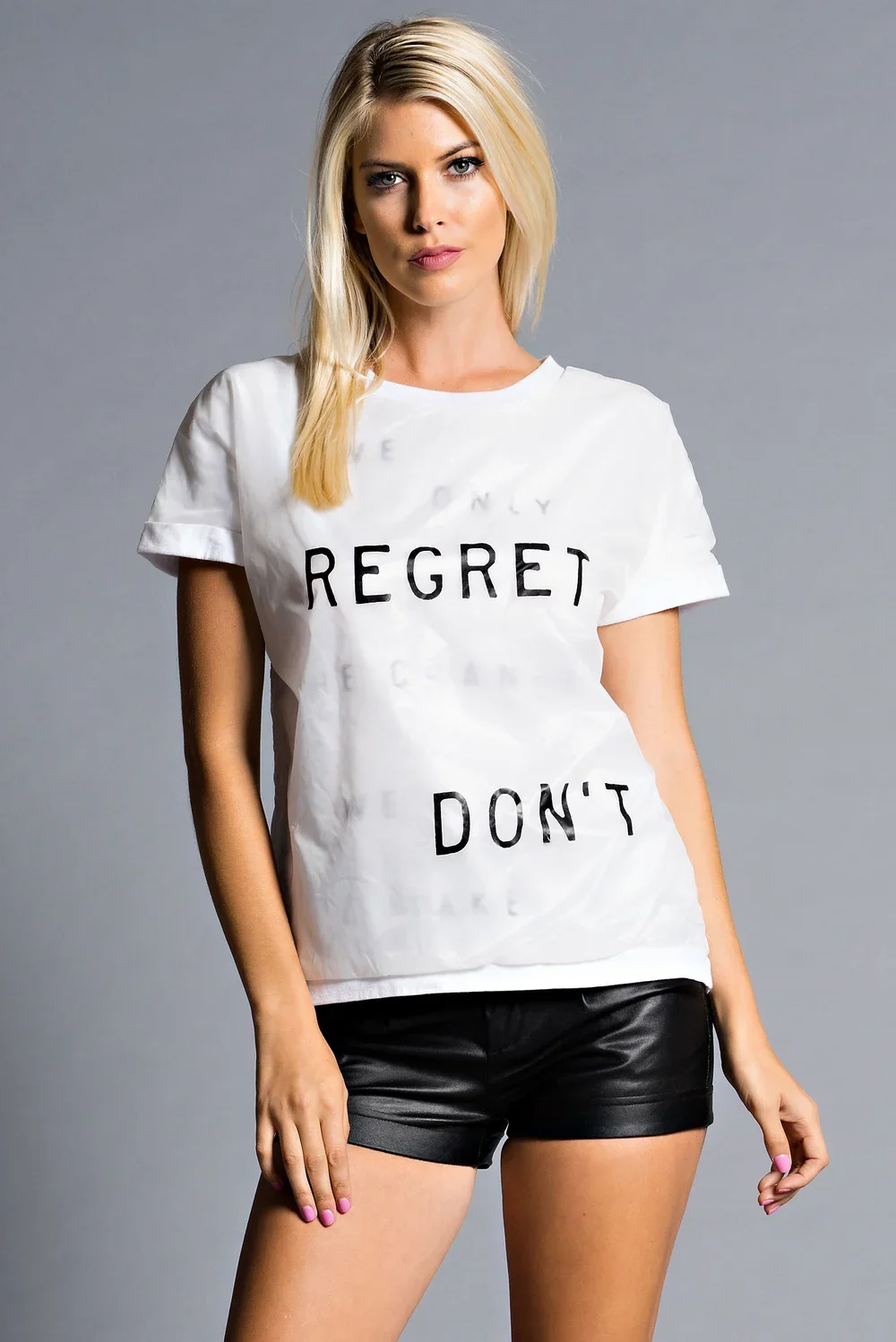 A young woman with blonde hair wearing a white T-shirt with the words "REGRET DON'T" printed in black and black shorts against a gray background.
