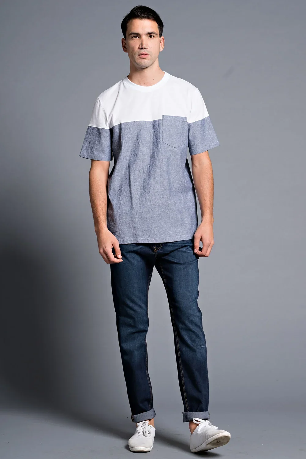 A young man with dark hair wearing a color-blocked T-shirt, dark jeans, and white sneakers posing against a gray background.