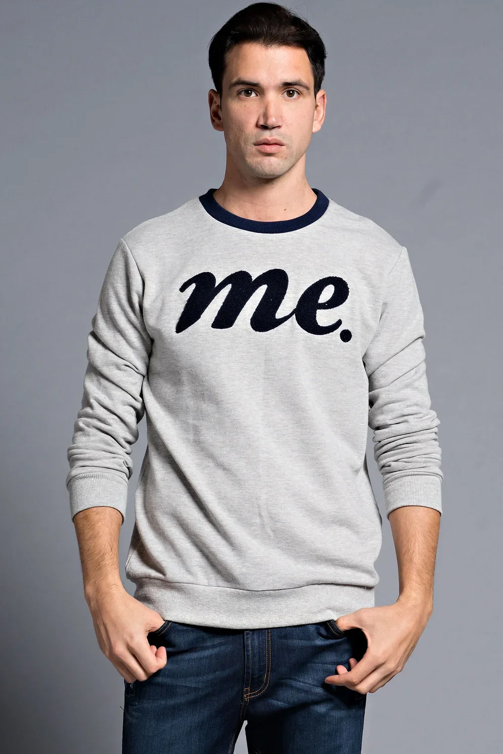 A man wearing a gray sweatshirt with the word "me." written on it in black letters, standing against a gray background.