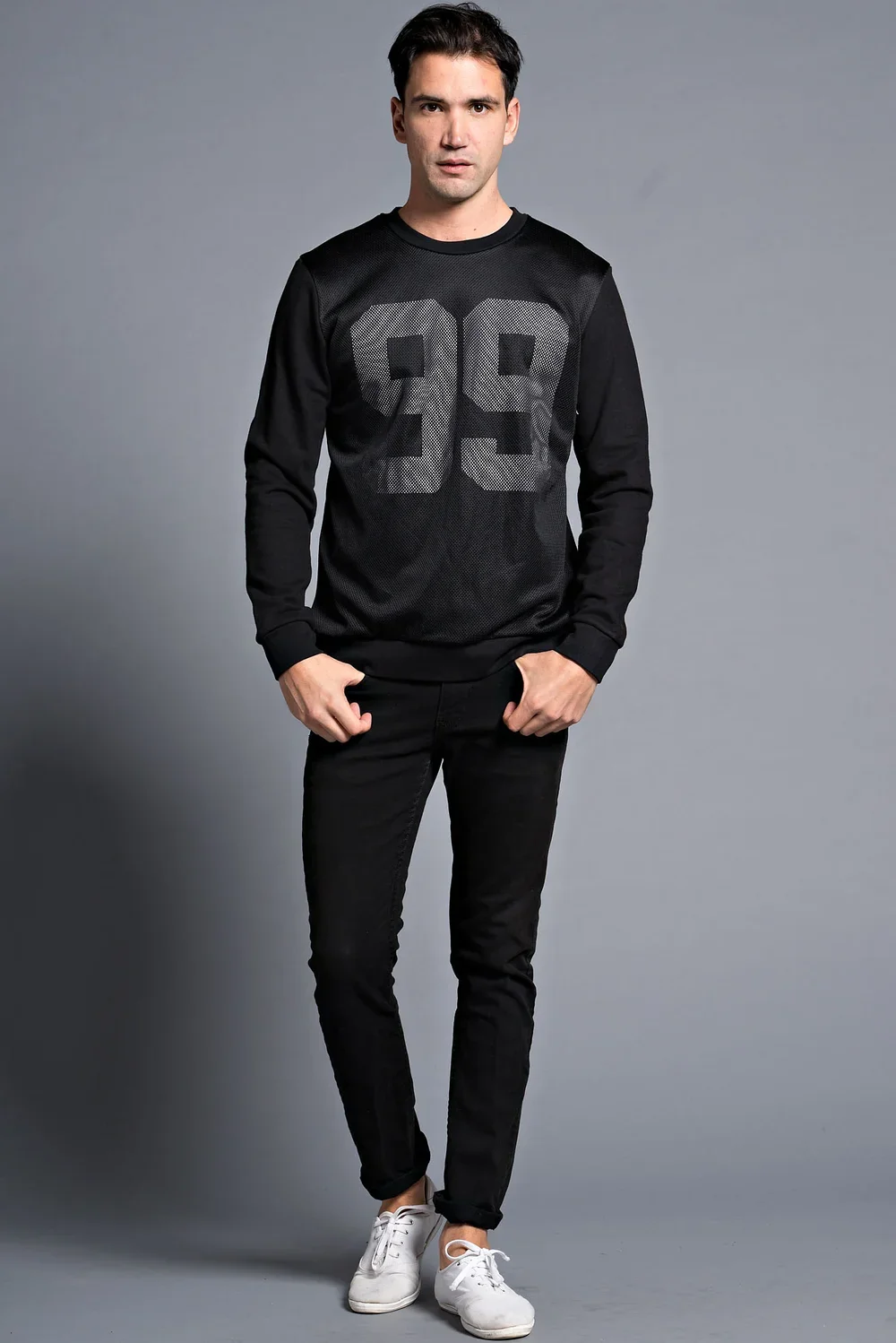 Young man wearing black sweatshirt with large gray number 99, black jeans, and white sneakers standing against a gray background.