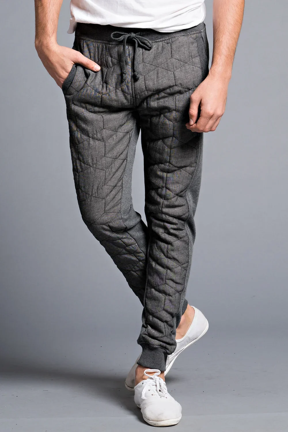 Close-up of a person wearing quilted gray sweatpants with a honeycomb pattern, a white t-shirt, and white sneakers, standing against a gray background with one hand in the pocket.