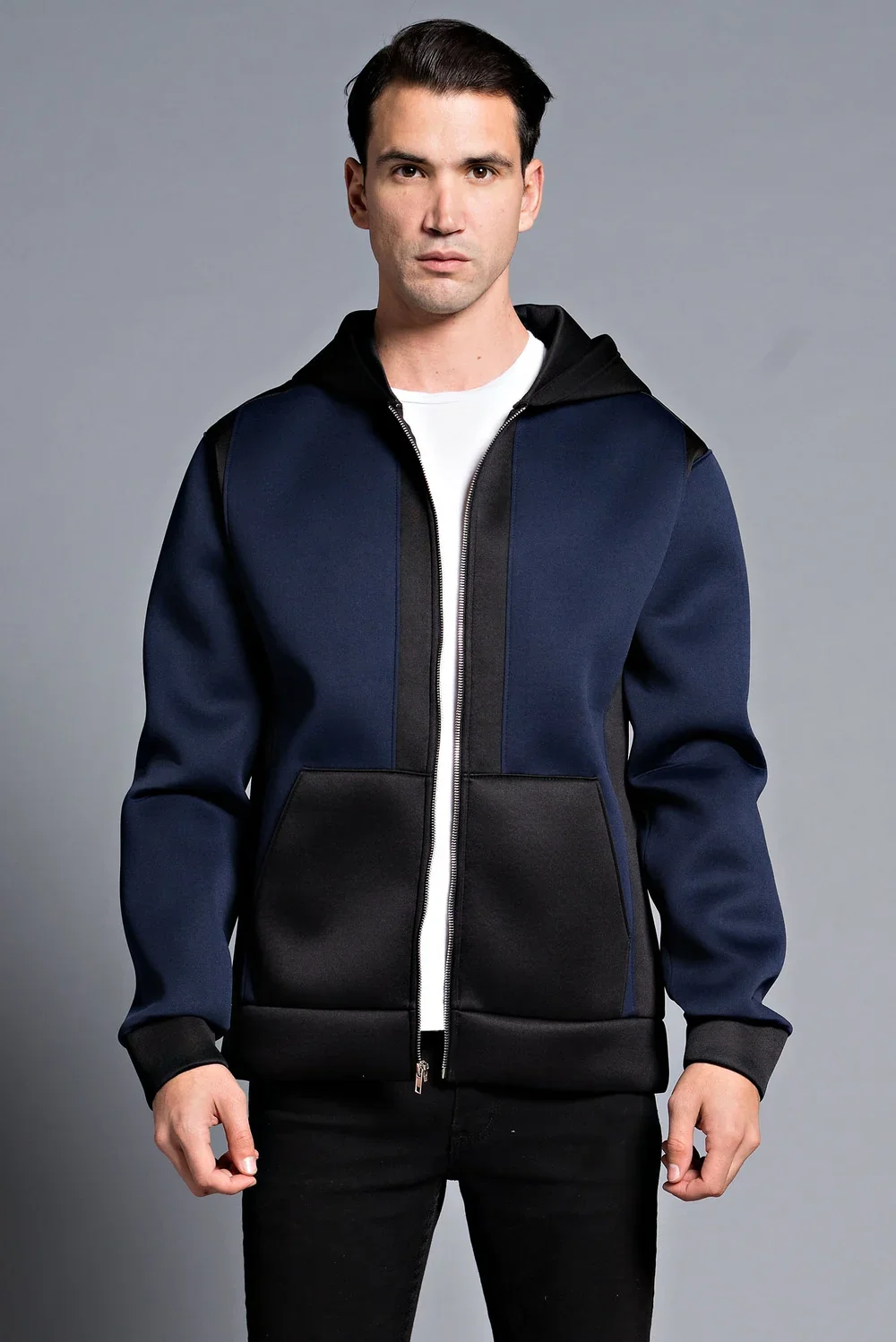 A man with short dark hair, wearing a navy zip-up hoodie with black accents and a white T-shirt underneath, standing against a plain gray background.