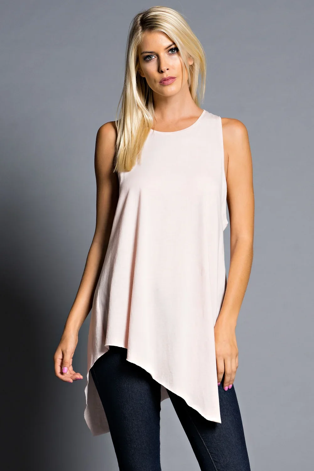 A blonde woman wearing a sleeveless white top and black pants standing against a grey background.