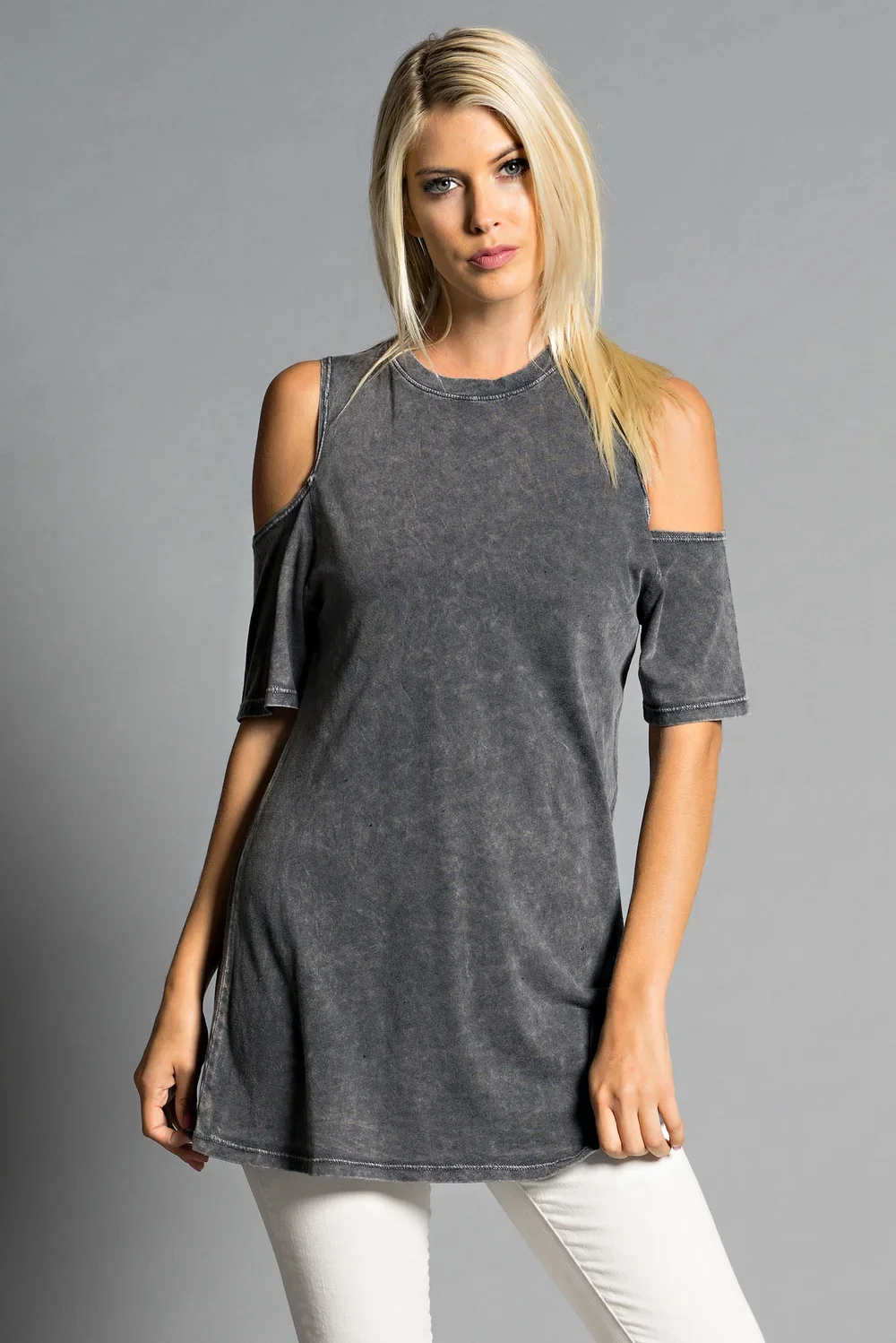 A young woman with long blonde hair wearing a grey cut-out shoulder t-shirt and white pants, standing against a neutral background.