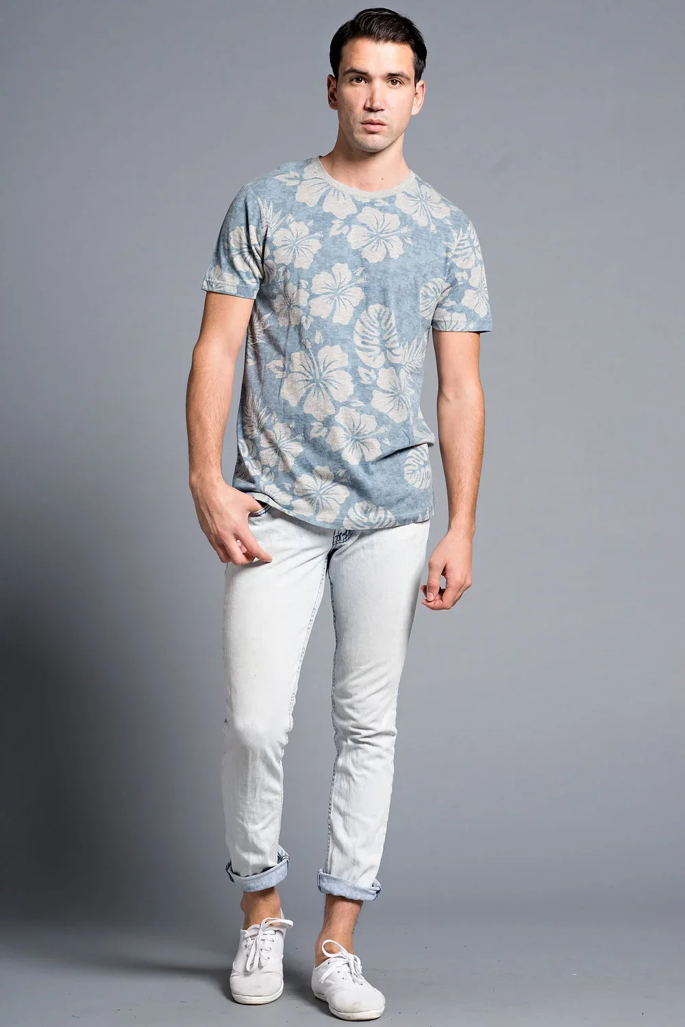 A young man with dark hair, wearing a blue floral T-shirt, white jeans with rolled cuffs, and white sneakers, standing against a gray background.
