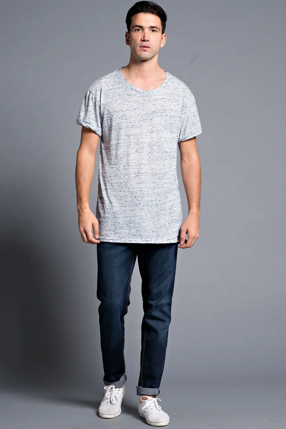 A young man standing against a plain gray background, wearing a casual gray t-shirt, dark jeans, and white sneakers.