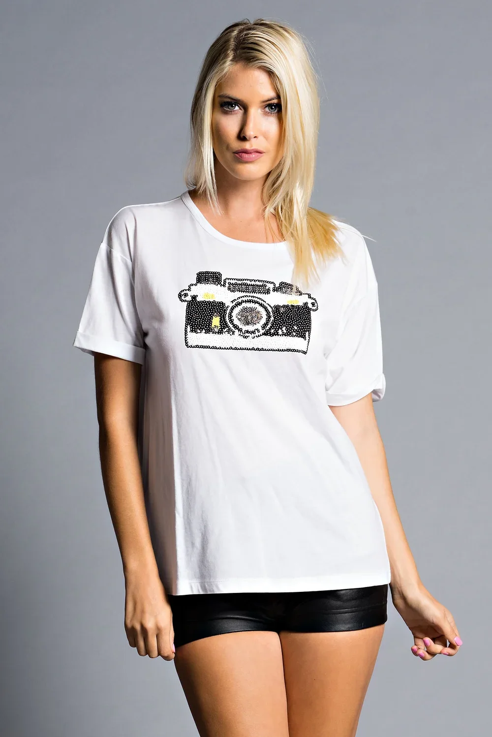 A woman with blonde hair wearing a white T-shirt with a sequin camera graphic and black shorts, standing against a plain gray background.