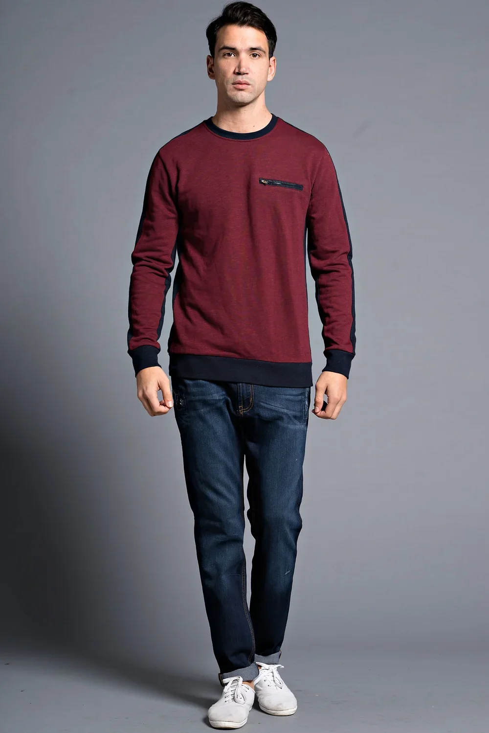 A young man with dark hair wearing a red and black long-sleeve sweatshirt with a zippered chest pocket, blue jeans, and white sneakers, standing against a plain gray background.