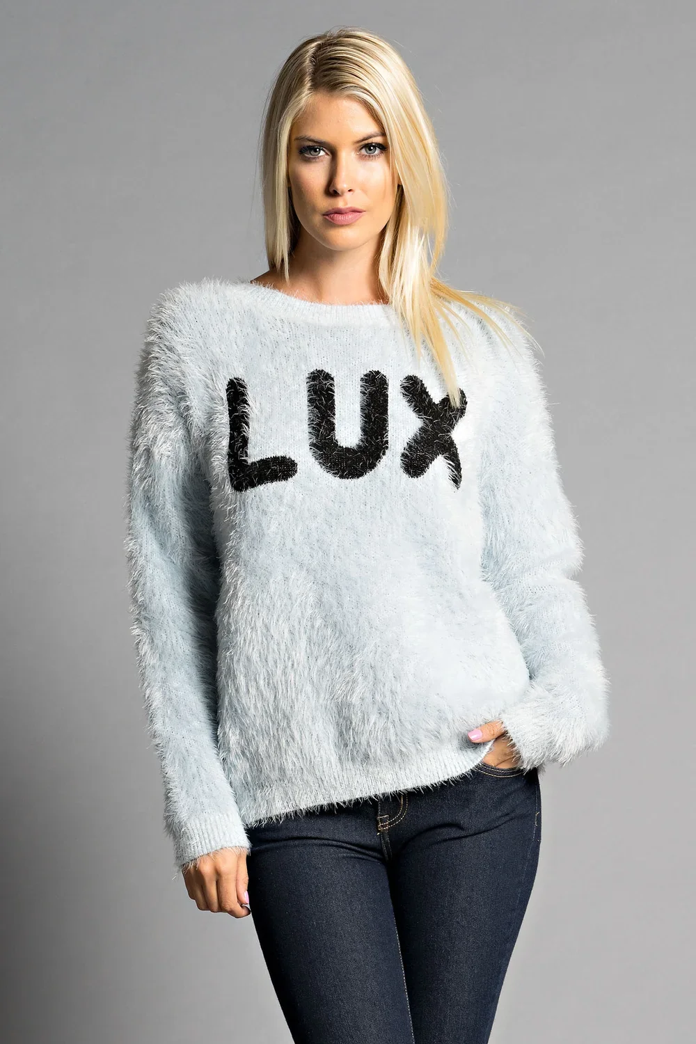 A young woman with blonde hair wearing a white fuzzy sweater with the word "LUX" in black on the front, and black jeans, standing against a plain gray background.