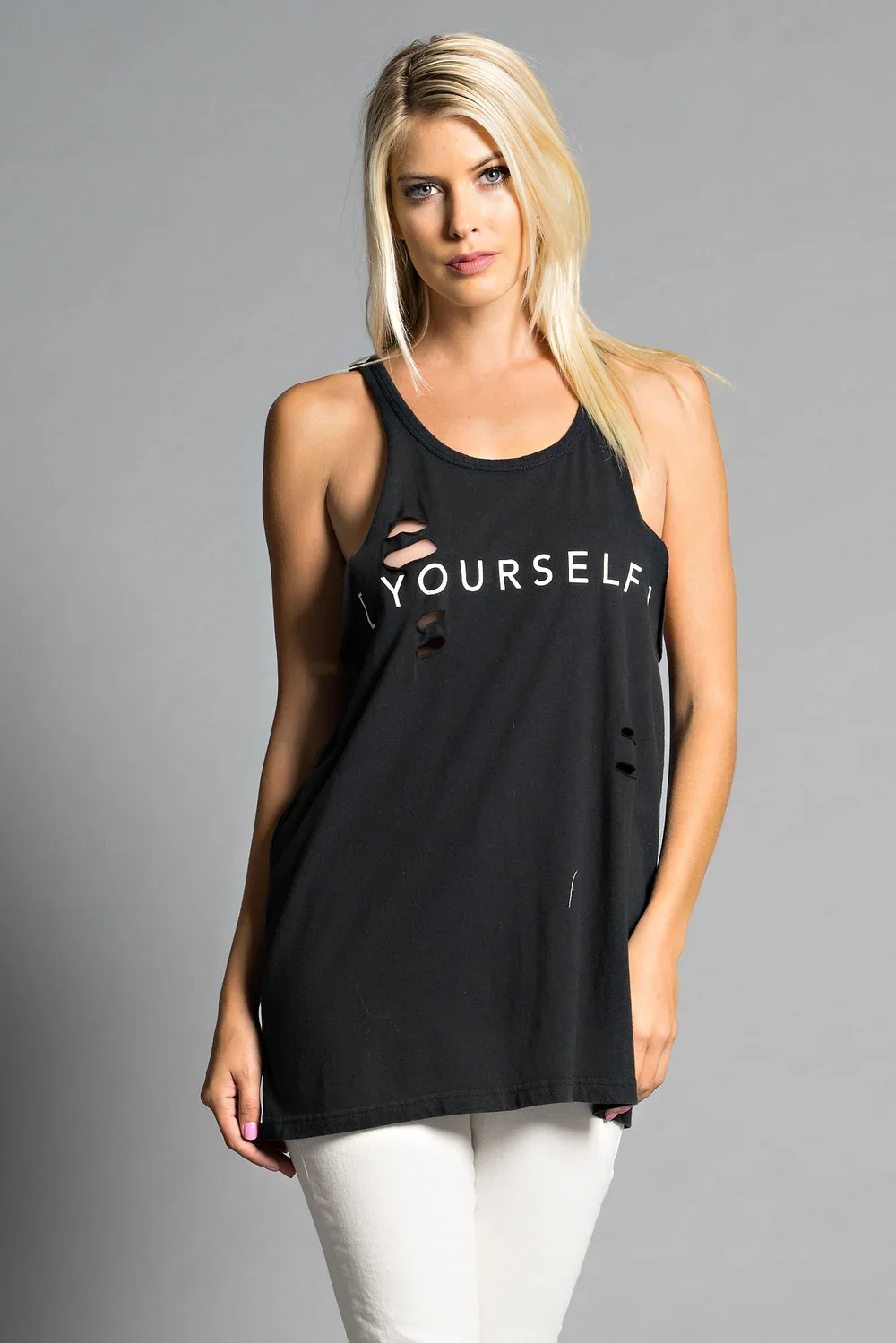 A woman with blonde hair wearing a distressed black tank top and white pants, standing against a gray background.