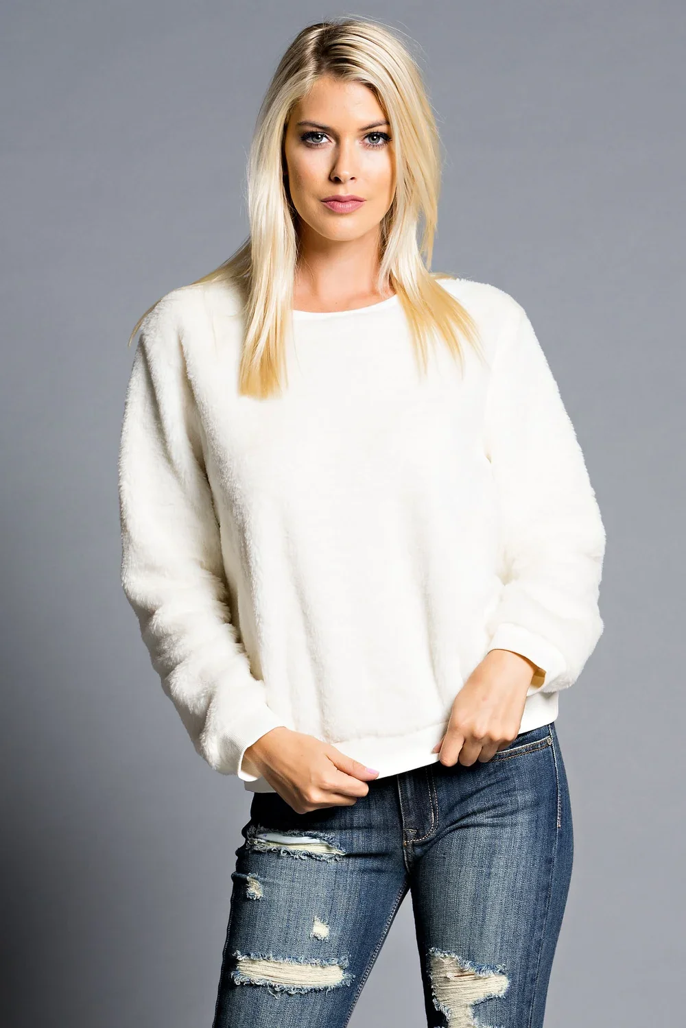 Woman with blonde hair wearing a cream sweater and distressed jeans against a gray background.