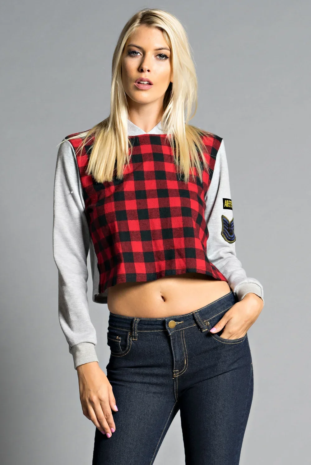 A young woman with blonde hair wears a cropped hoodie with gray sleeves and a red and black checkered front, standing against a gray background.