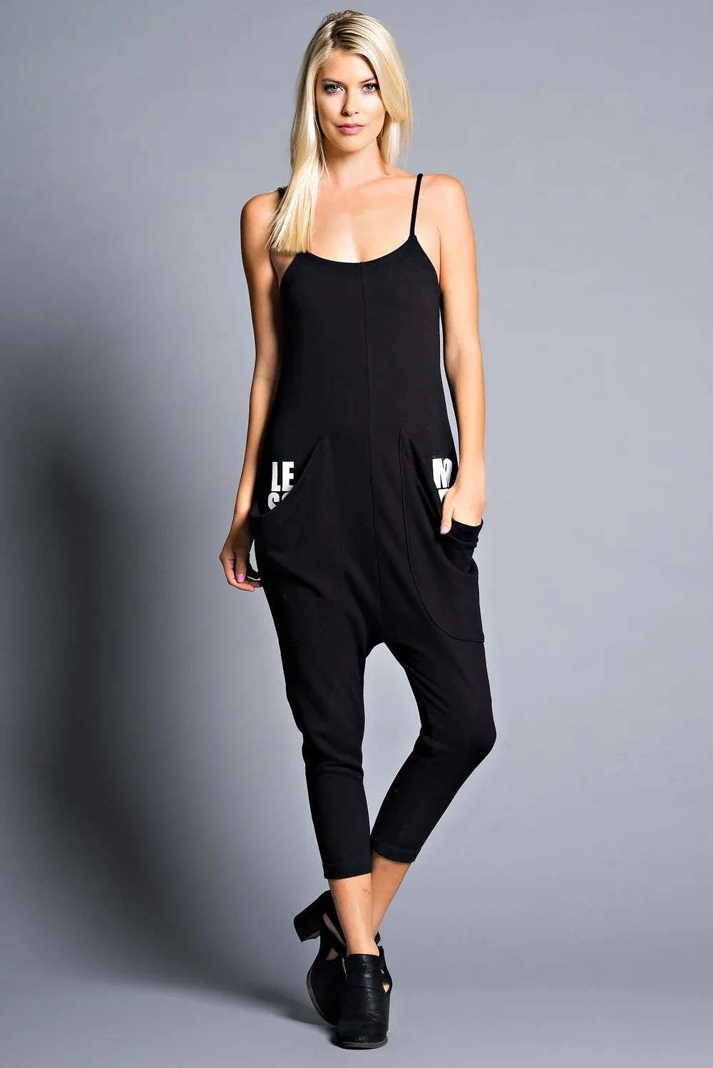A woman with blonde hair in a black sleeveless jumpsuit with pockets, standing against a plain gray background.