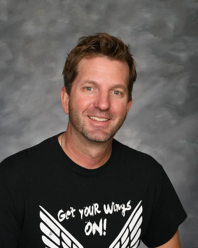 A smiling man with short brown hair and a beard, wearing a black T-shirt with the words "Get your wings on!" and wing graphics, standing against a gray backdrop.
