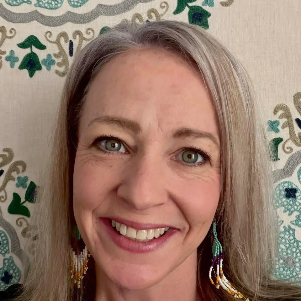 Close-up of a smiling woman with long blonde hair and earrings, standing in front of a decorative background with green, blue, and gold patterns.