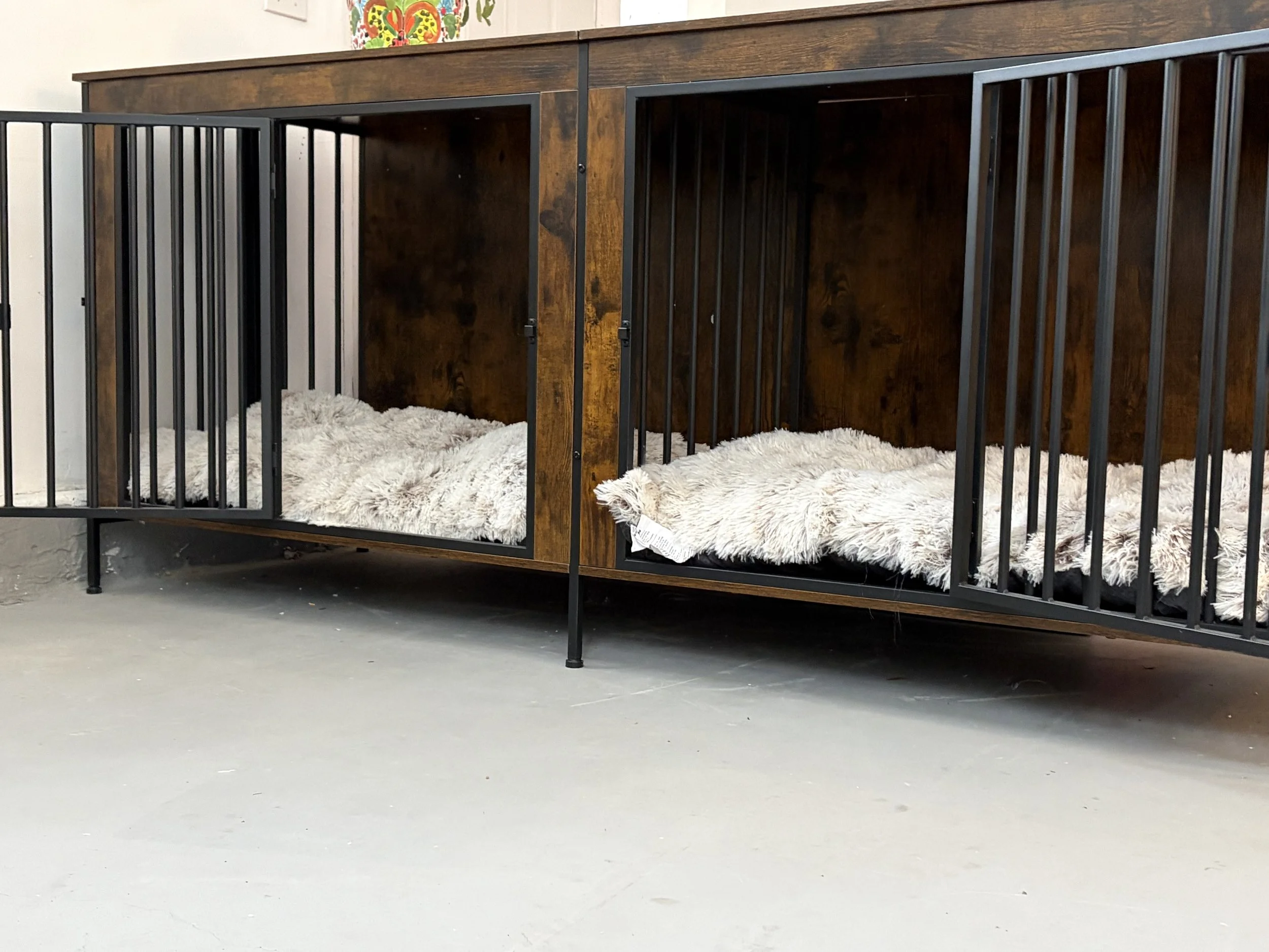 Two wooden and black metal pet crates with fluffy bedding inside, placed on a concrete floor against a wall with colorful decorations.
