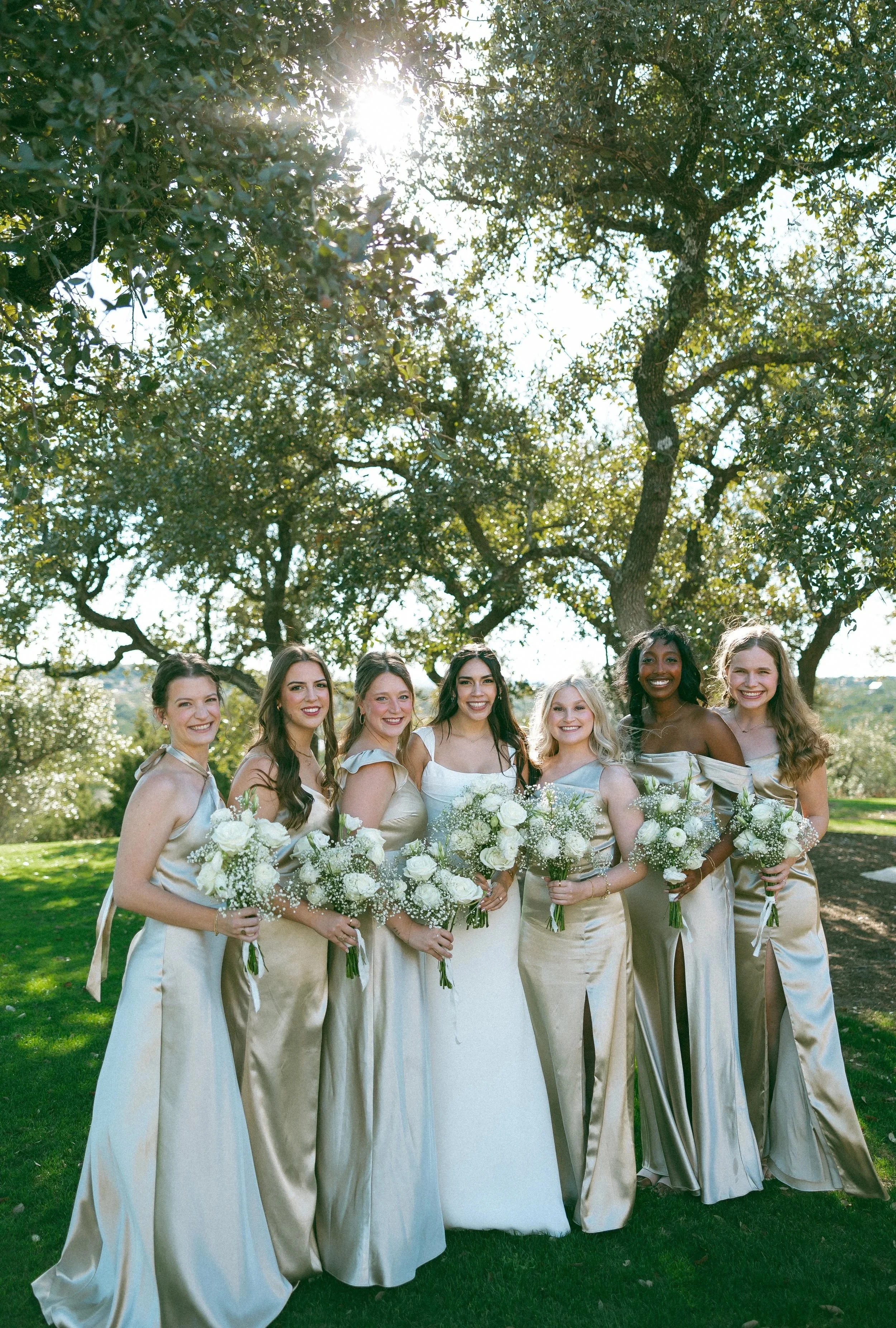 A group of eight women dressed in satin-colored bridesmaid dresses holding bouquets of white flowers, standing outdoors on a sunny day beneath large leafy trees.
