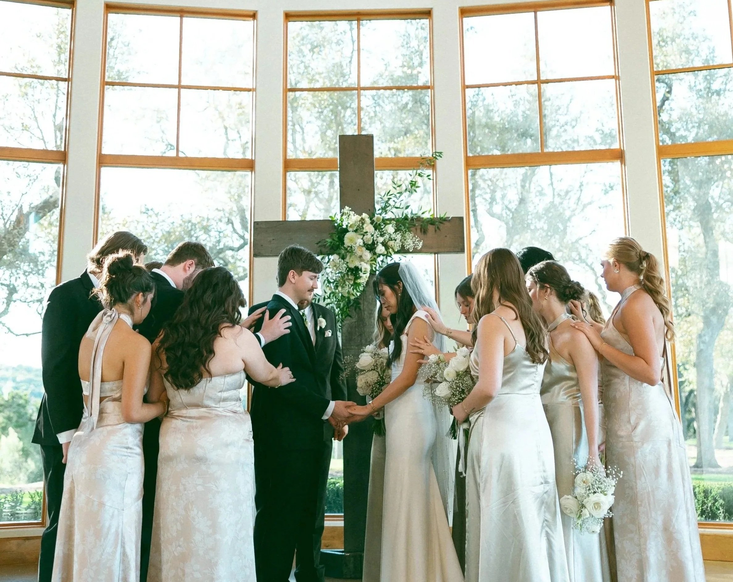 A wedding ceremony inside a church with a large cross and floral arrangements, with wedding couple and guests praying with heads bowed.