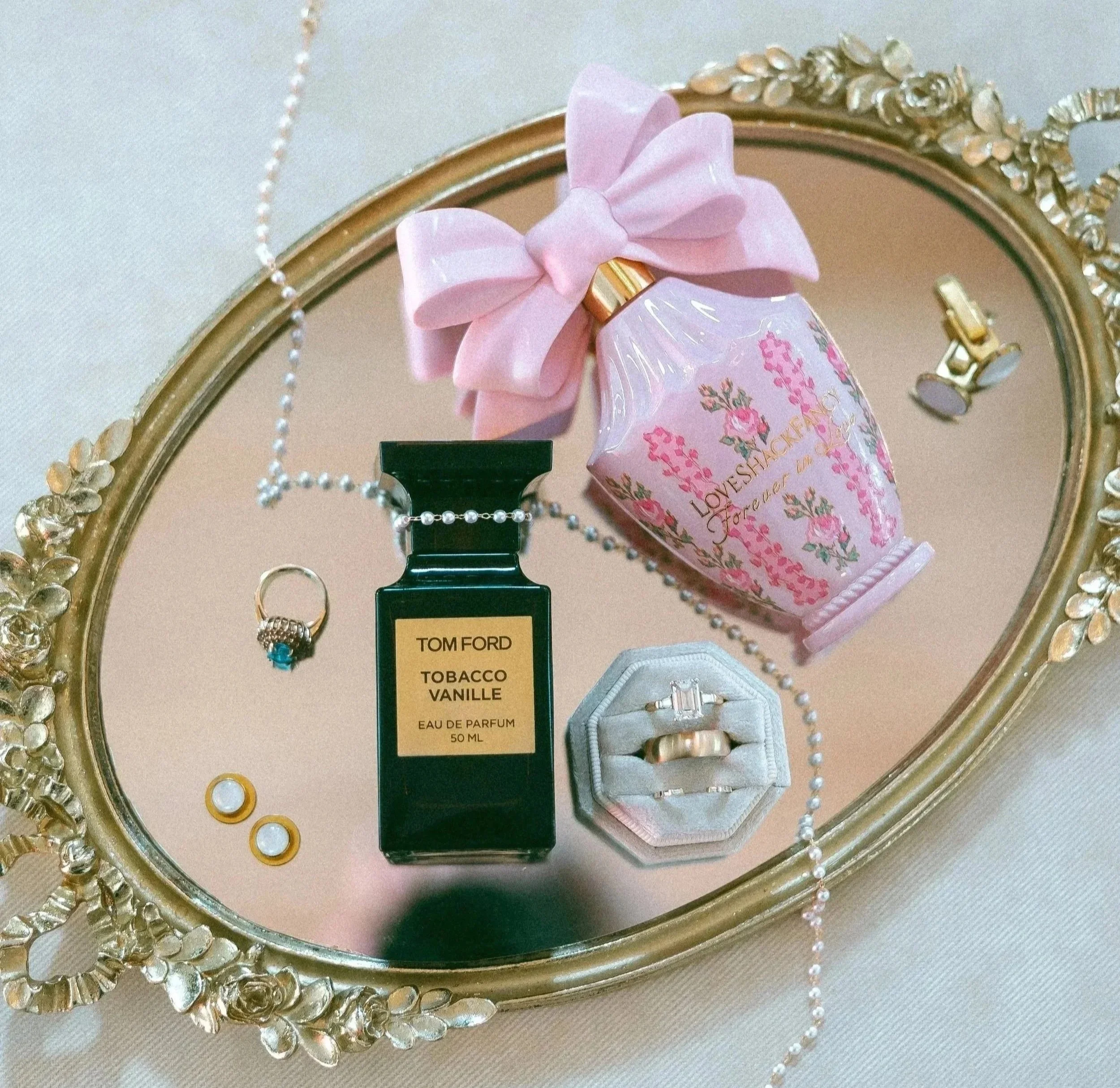 A decorative mirror with a gold ornate frame, displaying a pink perfume bottle with a bow and floral design, jewelry including rings, and pearl necklaces on a white surface.