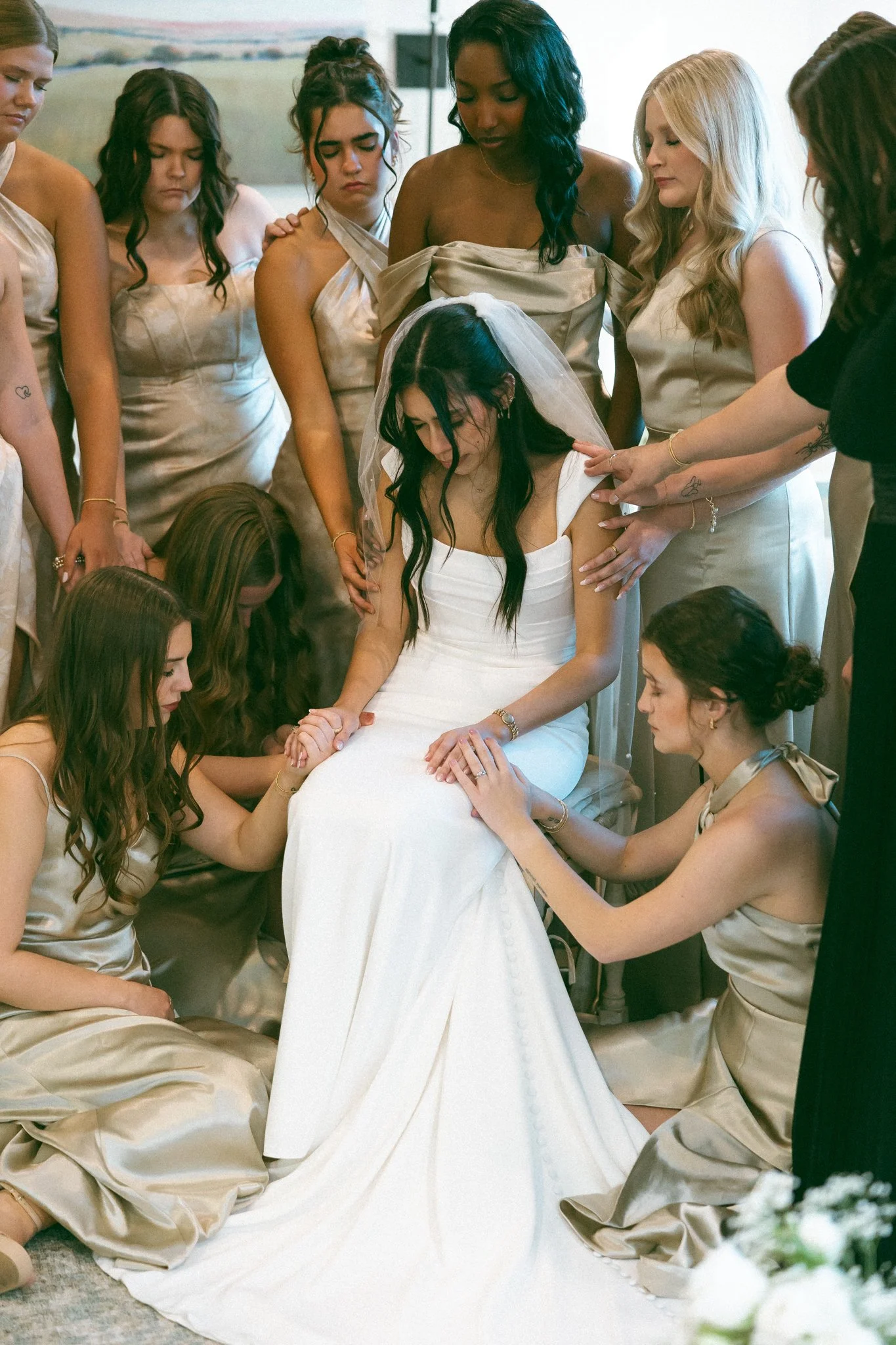 A bride in a white wedding dress with a veil is surrounded by women, who are offering prayer or blessings and holding her hands during a wedding ceremony.