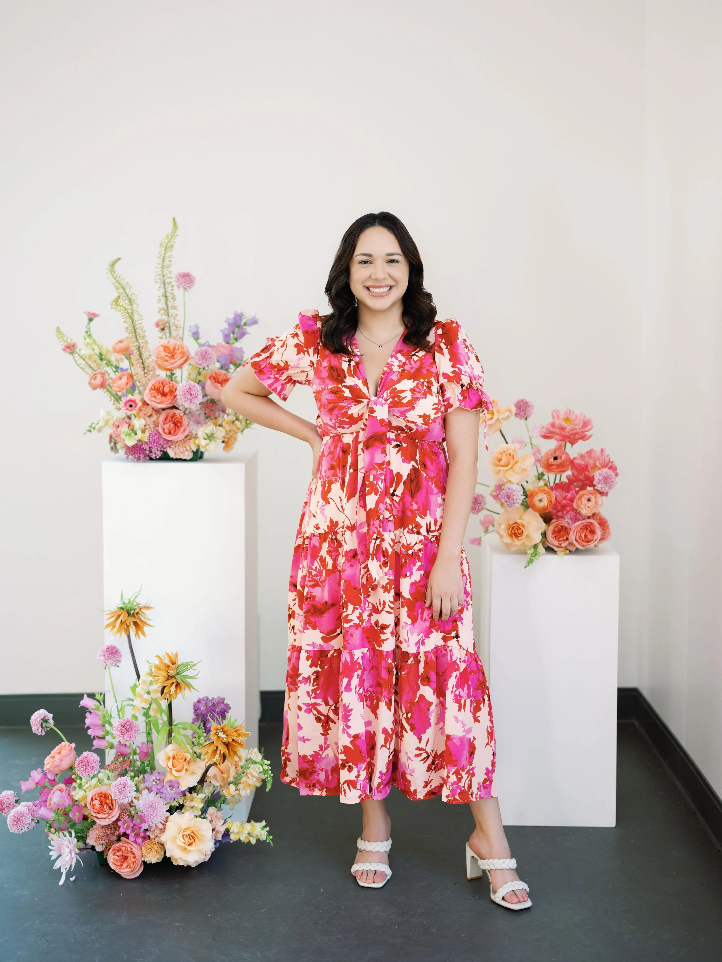 Headshot of founder of The Velvet Vow, a luxury wedding planner based in Charlotte, North Carolina serving. Woman with dark hair smiling and posing next to colorful flower arrangements, wearing a pink and red floral dress and white high-heeled shoes.