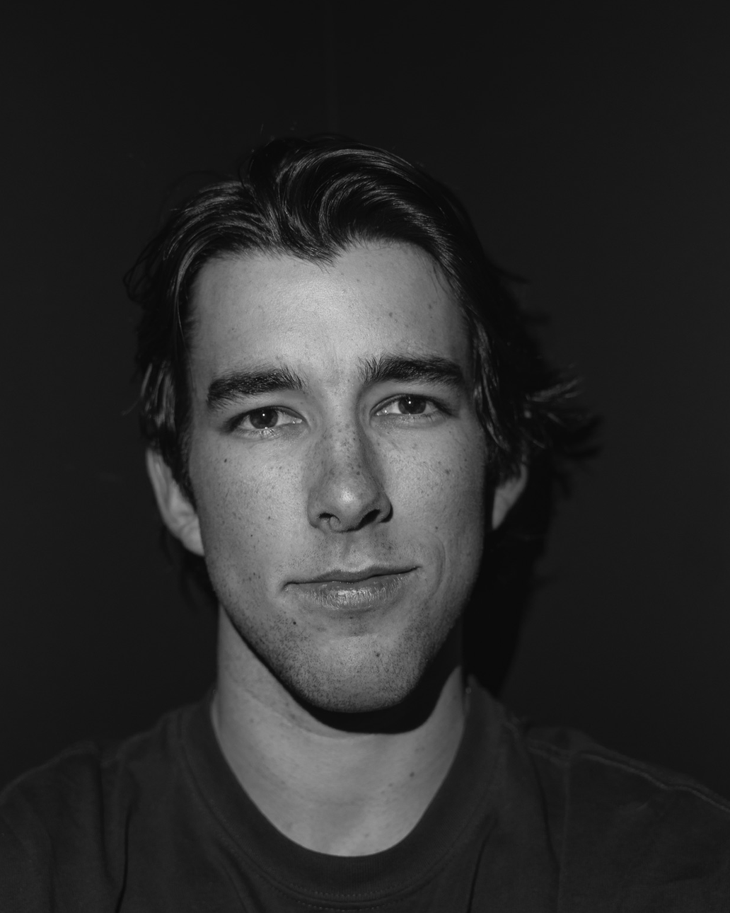 Black and white portrait of a young man with dark hair, slight smile, and freckles, wearing a dark t-shirt.