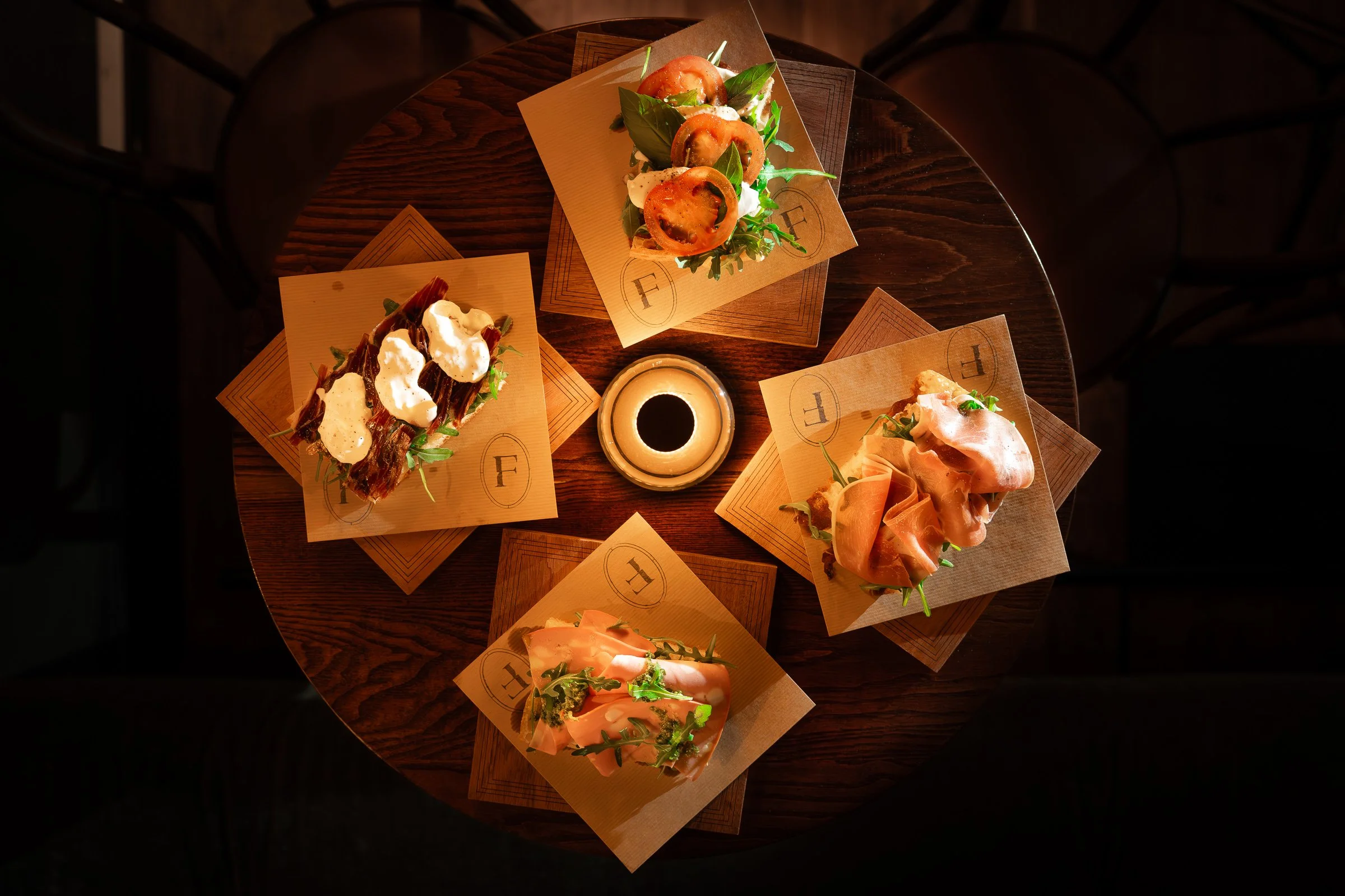 Four gourmet open-faced sandwiches arranged on a round wooden table with a small cup of dark sauce or coffee in the center. Each sandwich is on a different square placemat, topped with ingredients like tomatoes, prosciutto, greens, and drizzled sauce