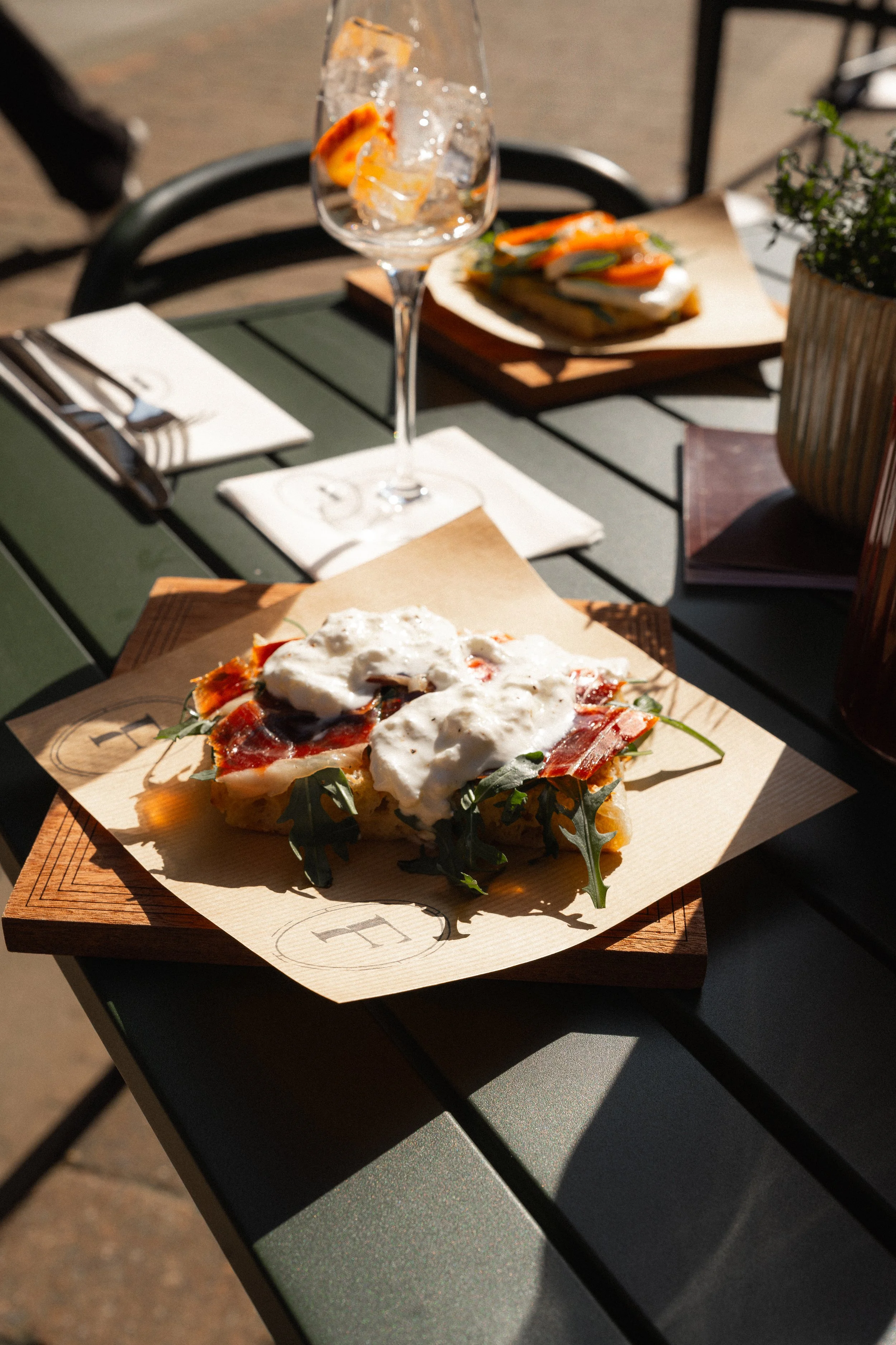 Close-up of a pizza topped with arugula, prosciutto, cheese, and cream, served on a wooden board at an outdoor cafe table with a glass of water and a plate of open-faced sandwich in the background.