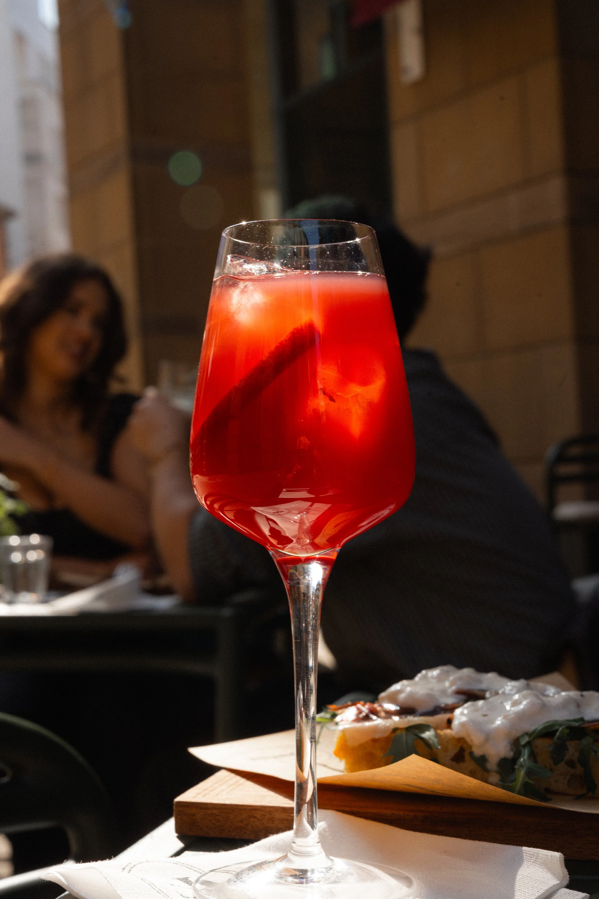 A tall glass of red cocktail with ice and a straw, placed on a table at a restaurant during daylight.