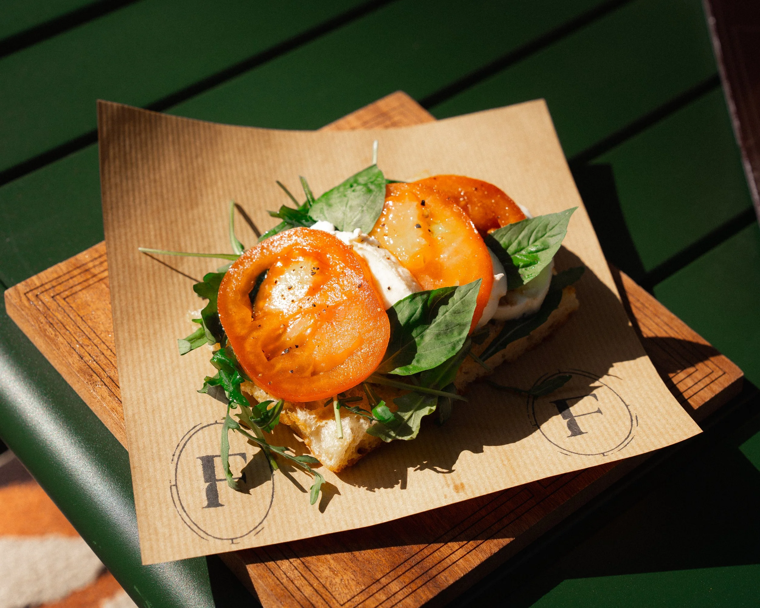 Open-faced sandwich with tomato slices, fresh herbs, and cheese on a piece of bread on a wooden serving board.