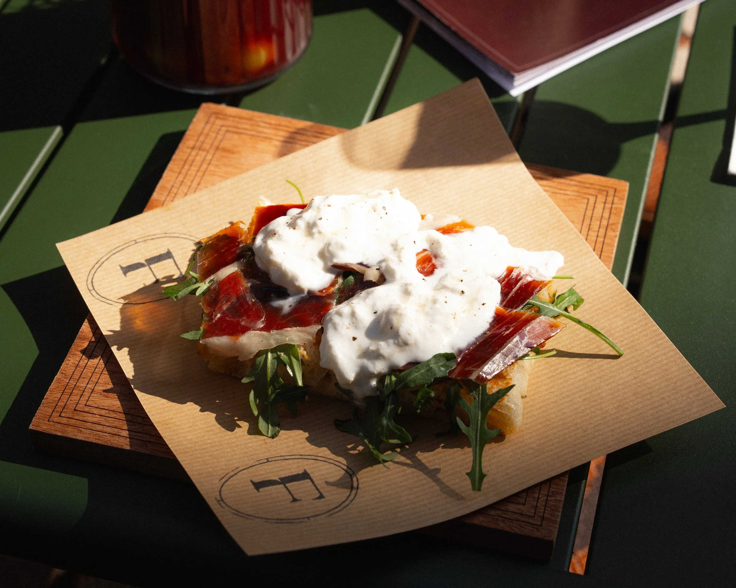 Open-faced sandwich with prosciutto, arugula, mozzarella cheese, and tomato sauce on bread, served on a paper-lined wooden board