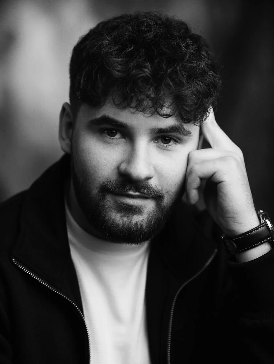 Black and white portrait of a young man with curly hair, beard, wearing a dark jacket and a white shirt, looking directly at the camera.
