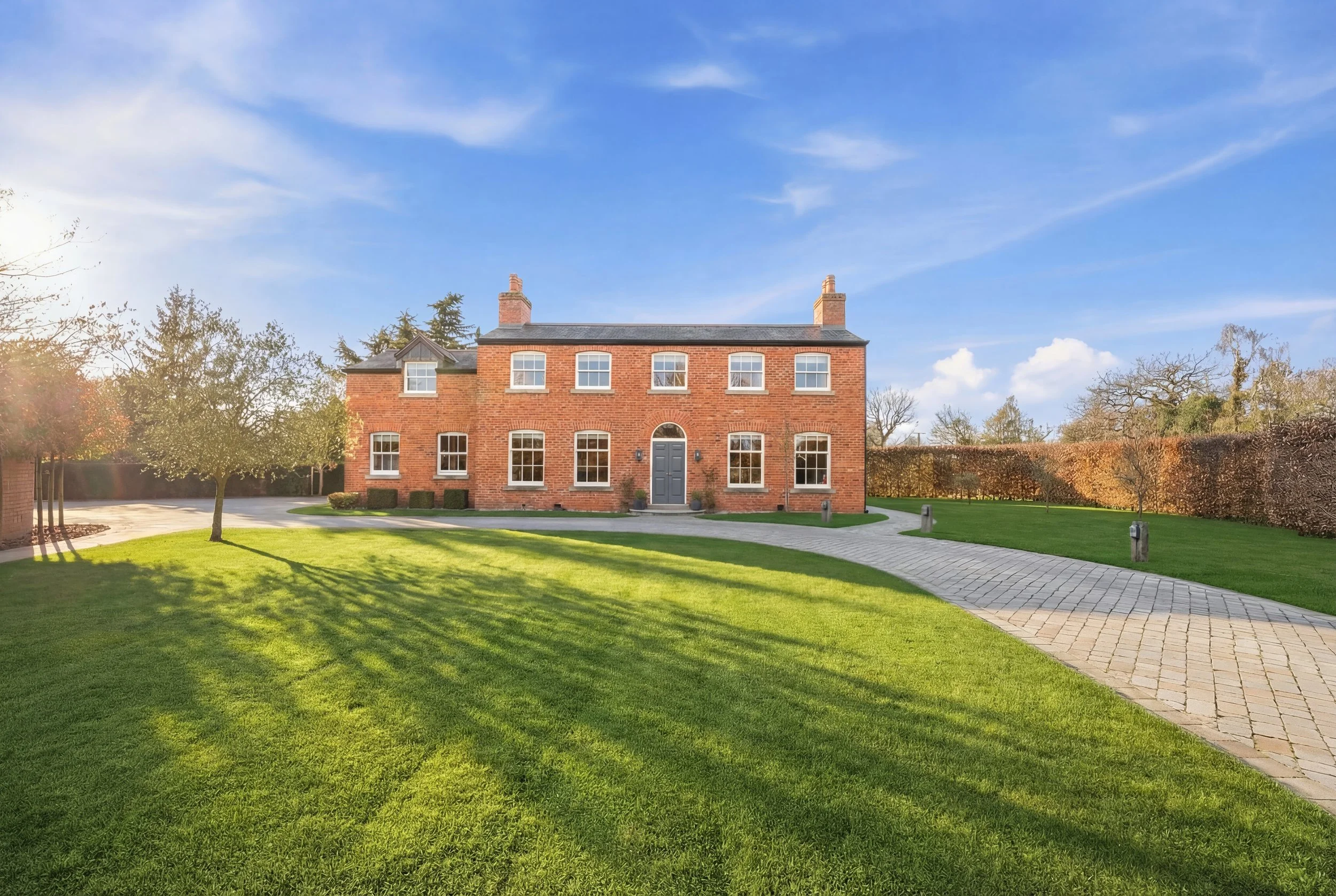 A large brick house with a well-maintained lawn, a curved paved driveway, and a blue sky with some clouds.