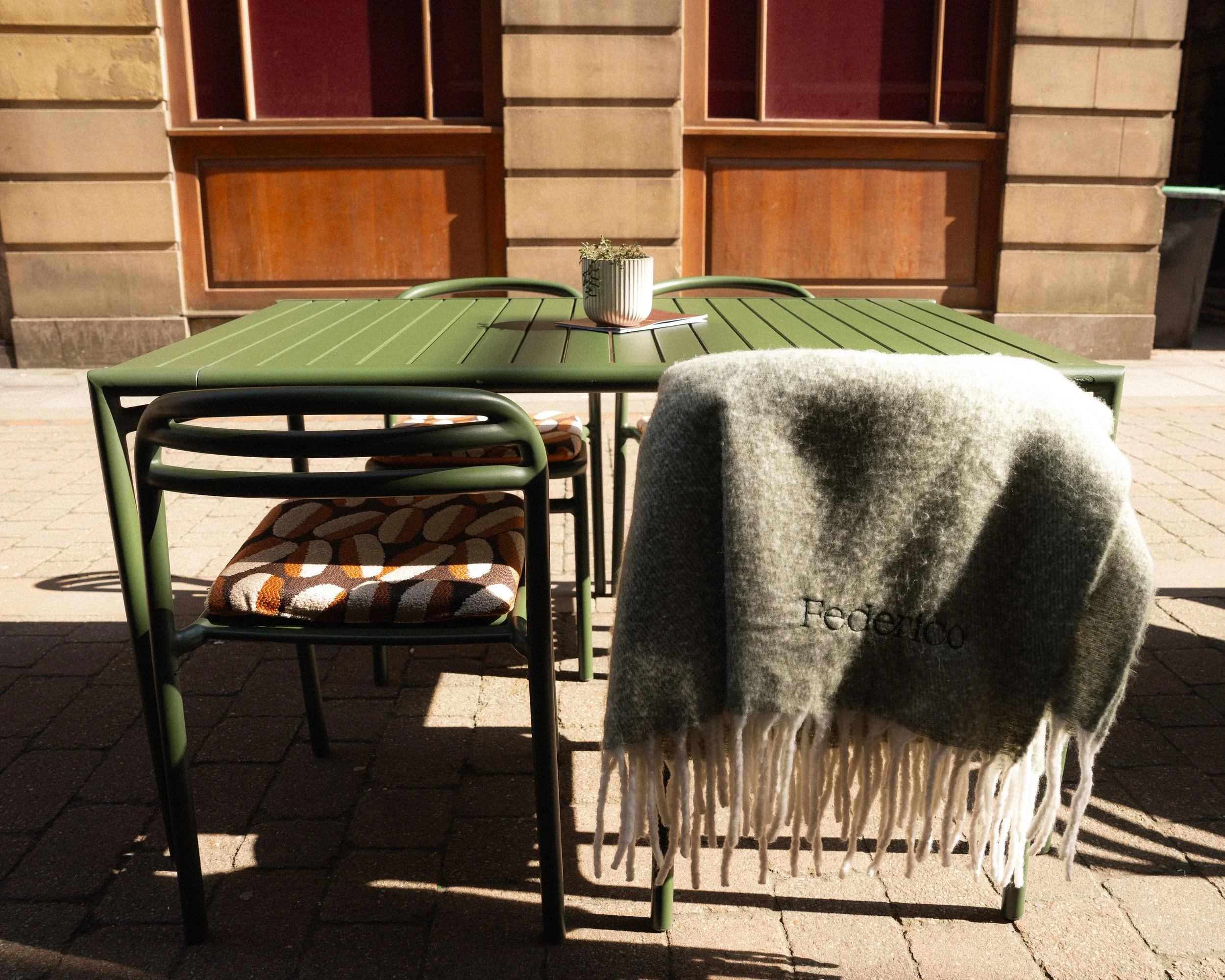 Outdoor green metal table with two matching chairs, a potted plant, and a gray blanket with the word 'Federico' hanging on the back of a chair, against a brick building background.