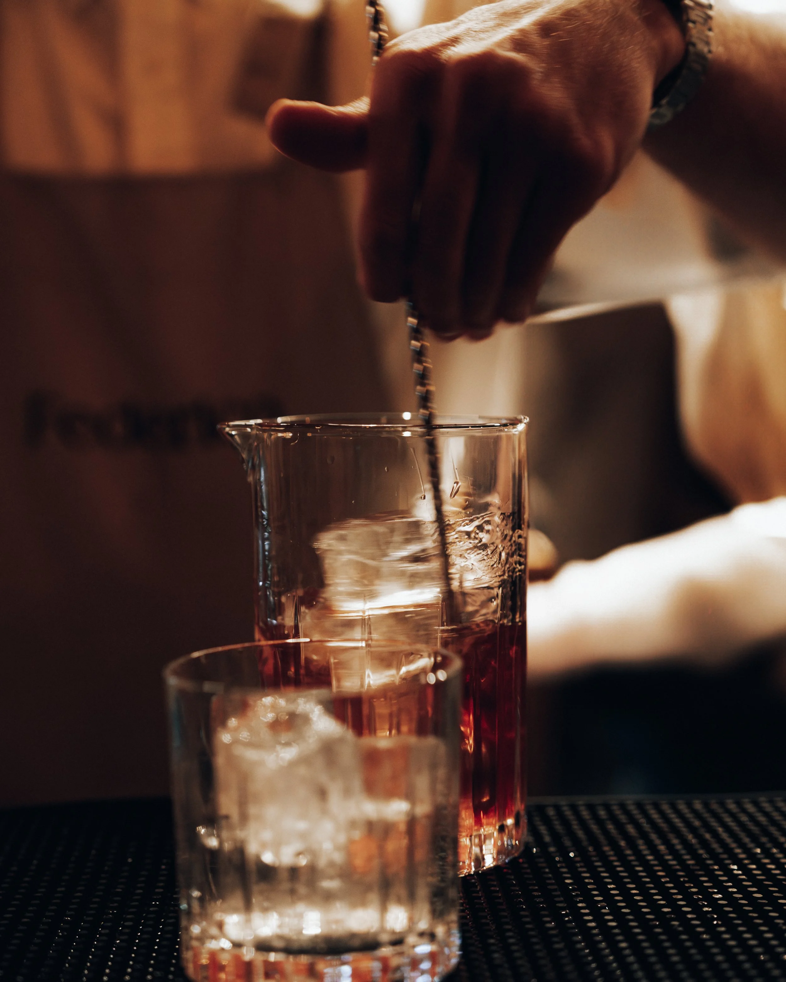 A person's hand pouring a red drink into a tall glass with ice, next to a smaller glass of ice on a bar counter.