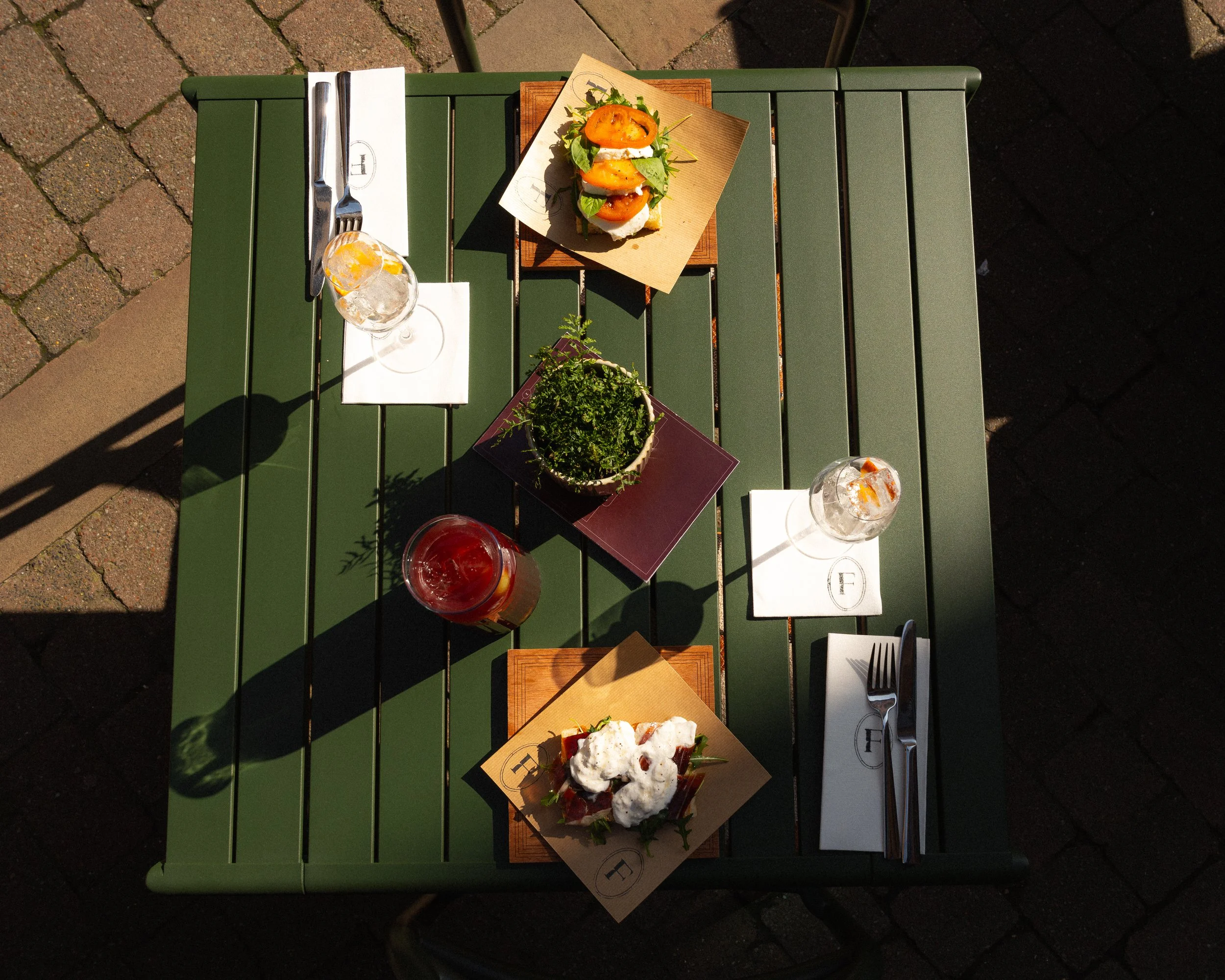 A green outdoor table set with appetizers, salads, drinks, and utensils at a sunny restaurant patio.