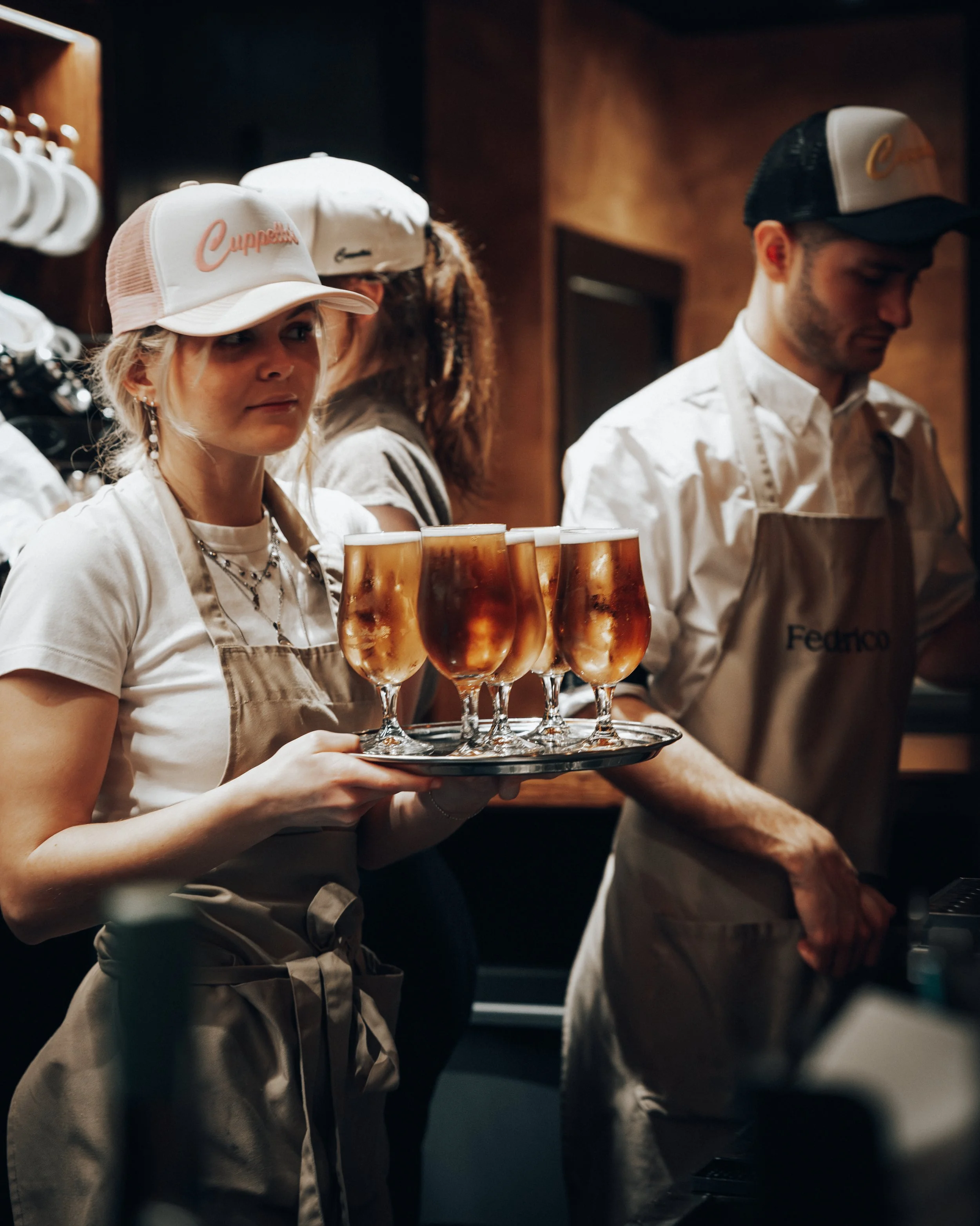 A woman holding a tray of six glasses filled with a dark amber beverage, likely beer, in a dimly lit bar or restaurant. She wears a beige apron and a white baseball cap.