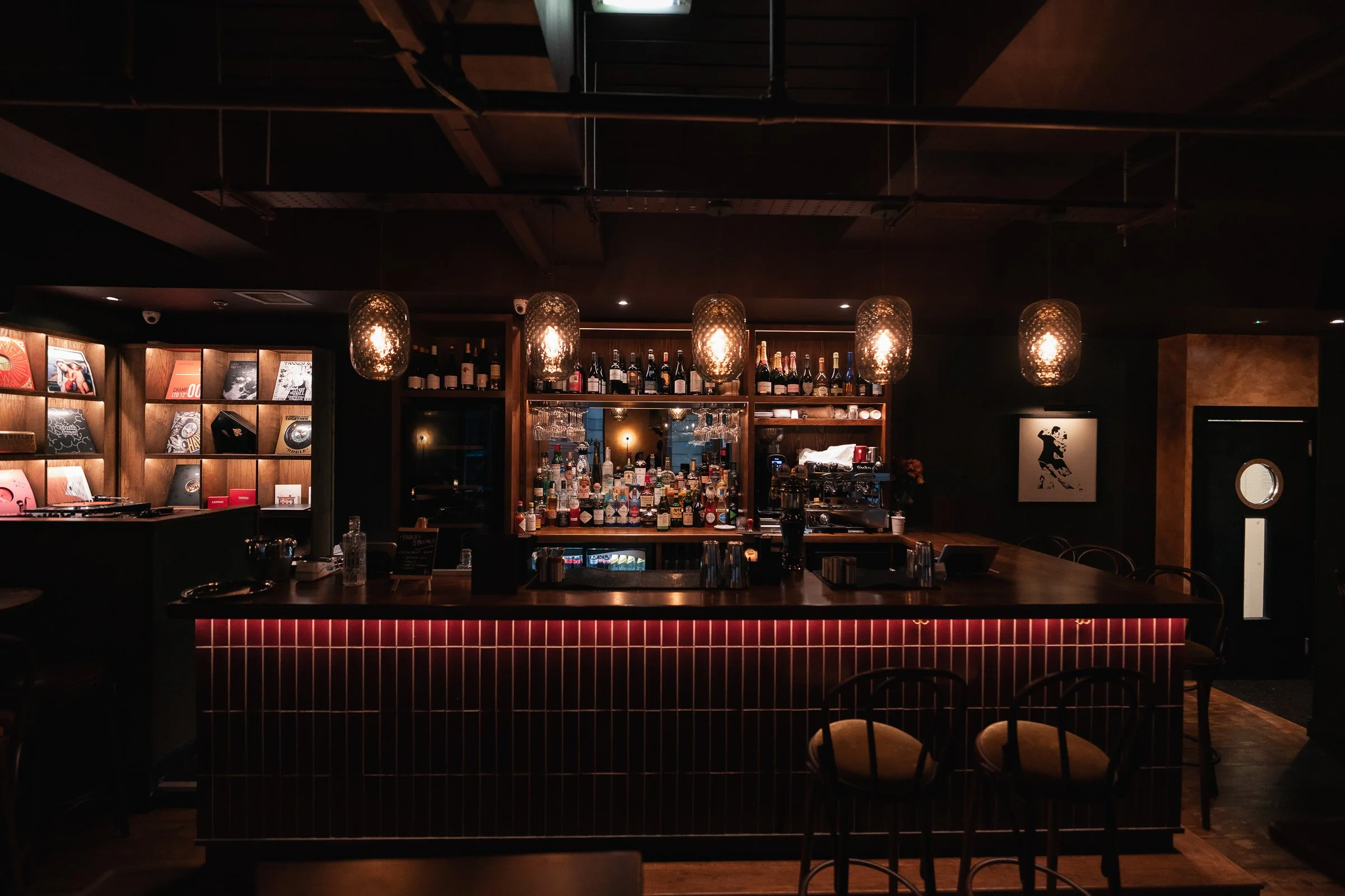  dimly lit bar with a backlit counter, shelves filled with assorted liquor bottles, hanging pendant lights with woven shades, and vinyl records on the wall to the left.