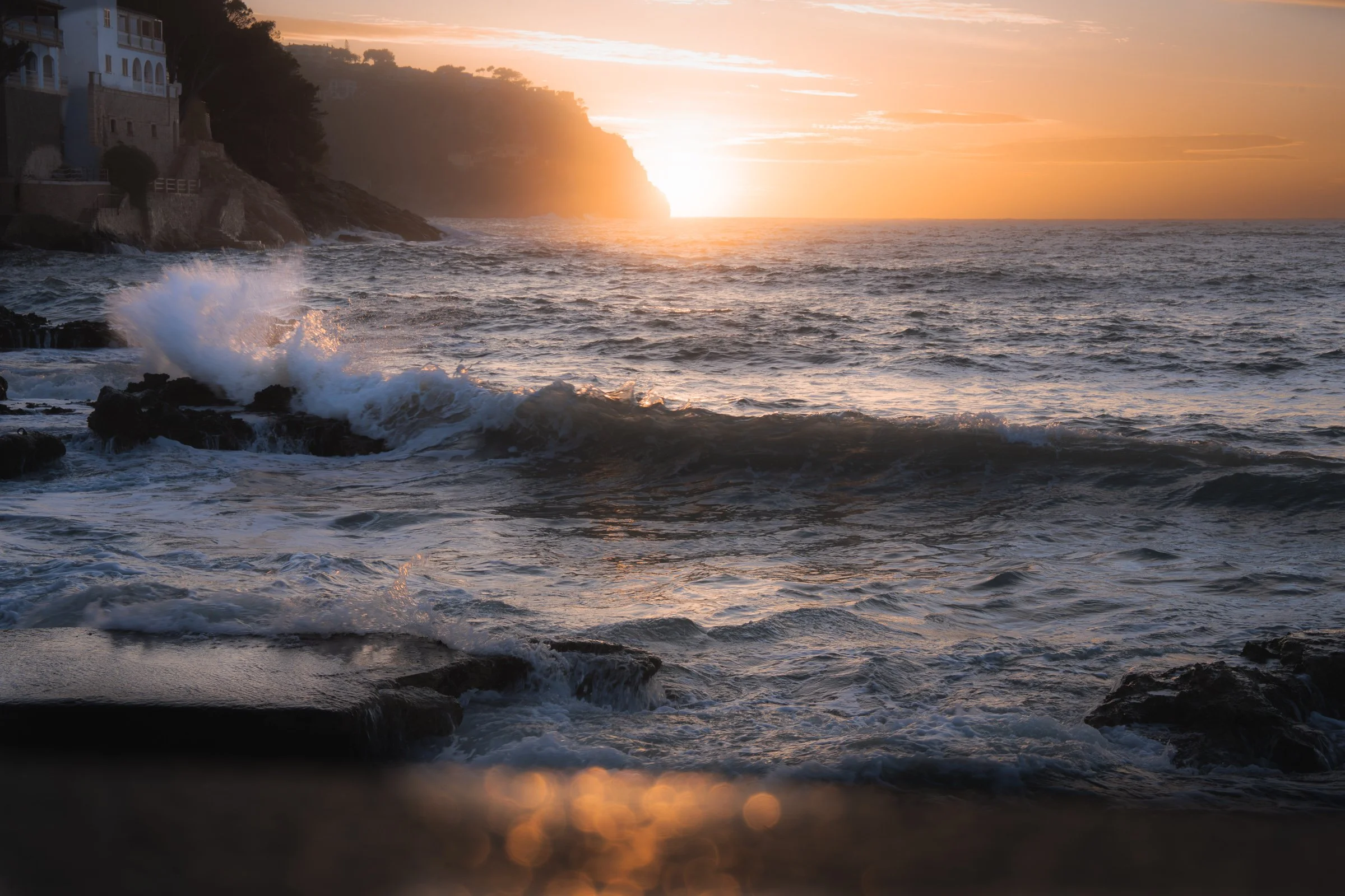 Sunset over the ocean with waves crashing against rocks along a coastal cliff, with buildings on the hillside to the left.