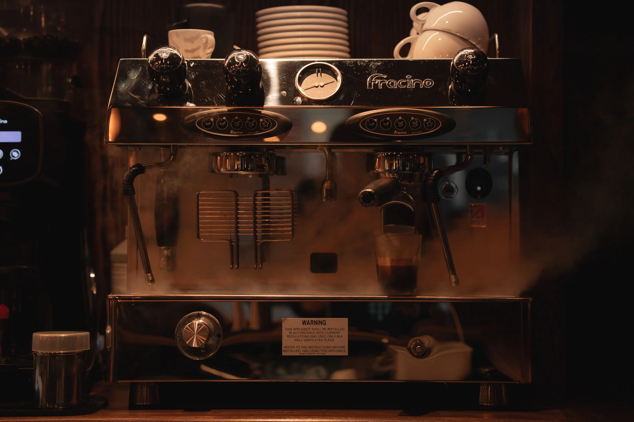 Behind a steaming espresso machine in a dimly lit setting, there are white cups and plates on top, with a few cups stacked. The machine has control buttons and a gauge, and a glass of espresso is collecting under one portafilter. A small warning labe