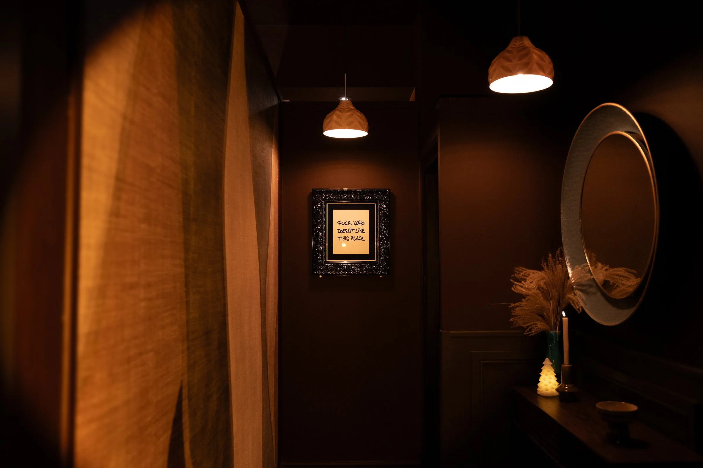 Dark hallway with warm lighting, framed artwork on the wall, a round mirror, a vase with dried plants, and a candle.
