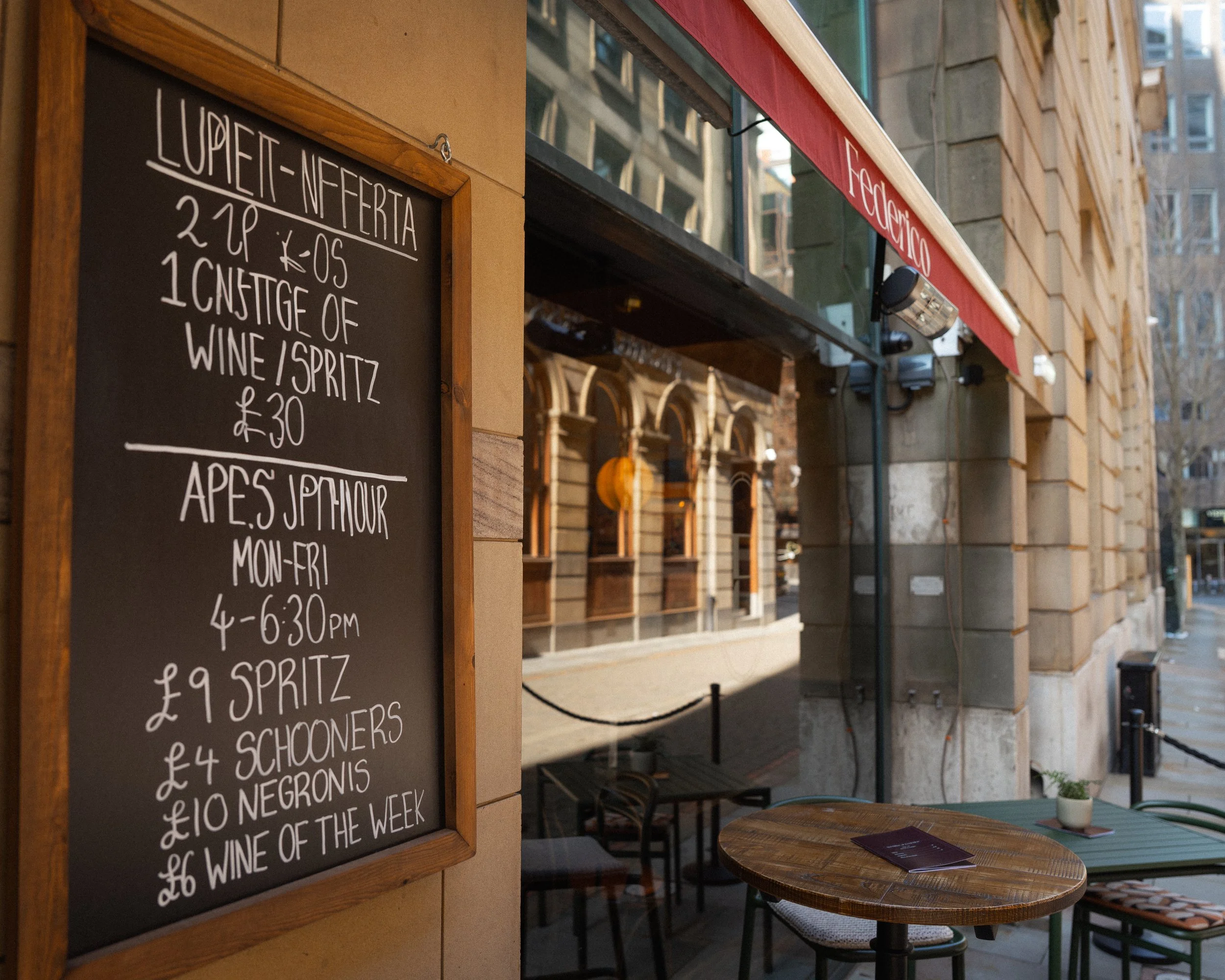 Outdoor sidewalk restaurant with chalkboard menu listing wine and spritz specials near a building with large windows and a red awning.