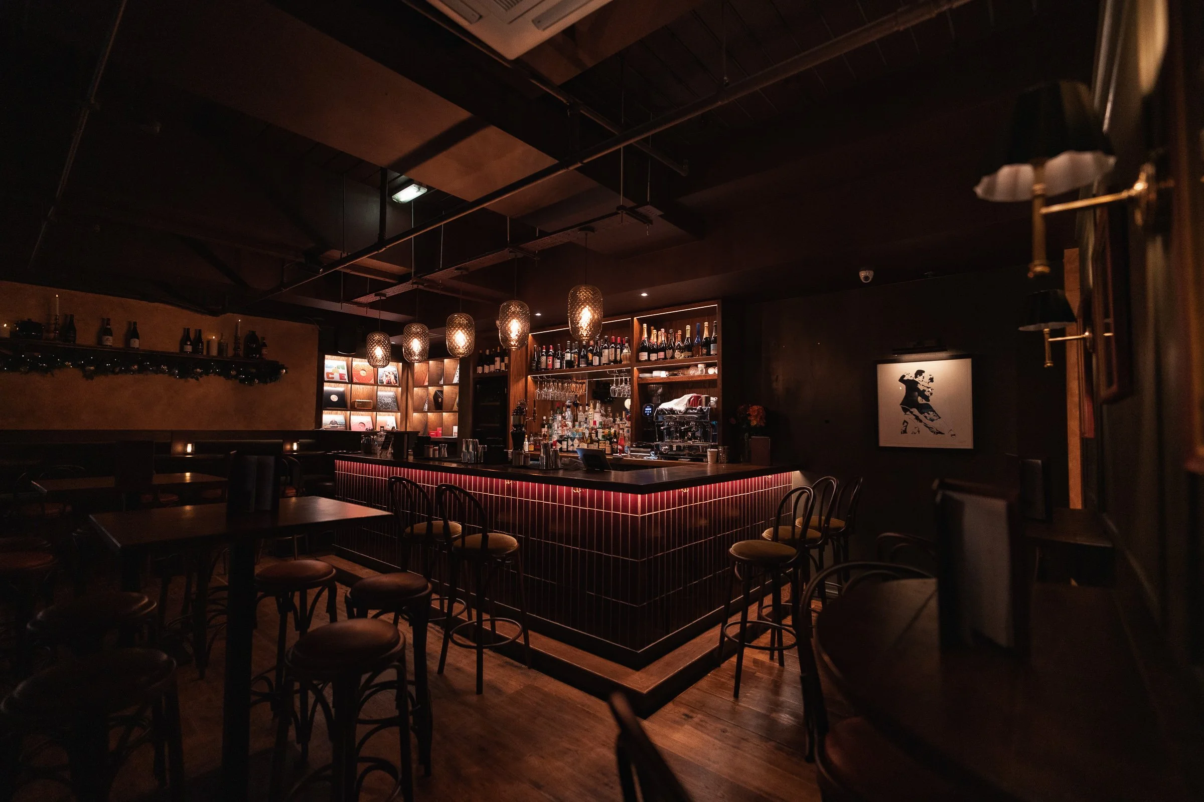Dimly lit bar with wooden shelves stocked with bottles and glasses, a central counter with a black countertop illuminated by hanging pendant lights, and several chairs and tables around.