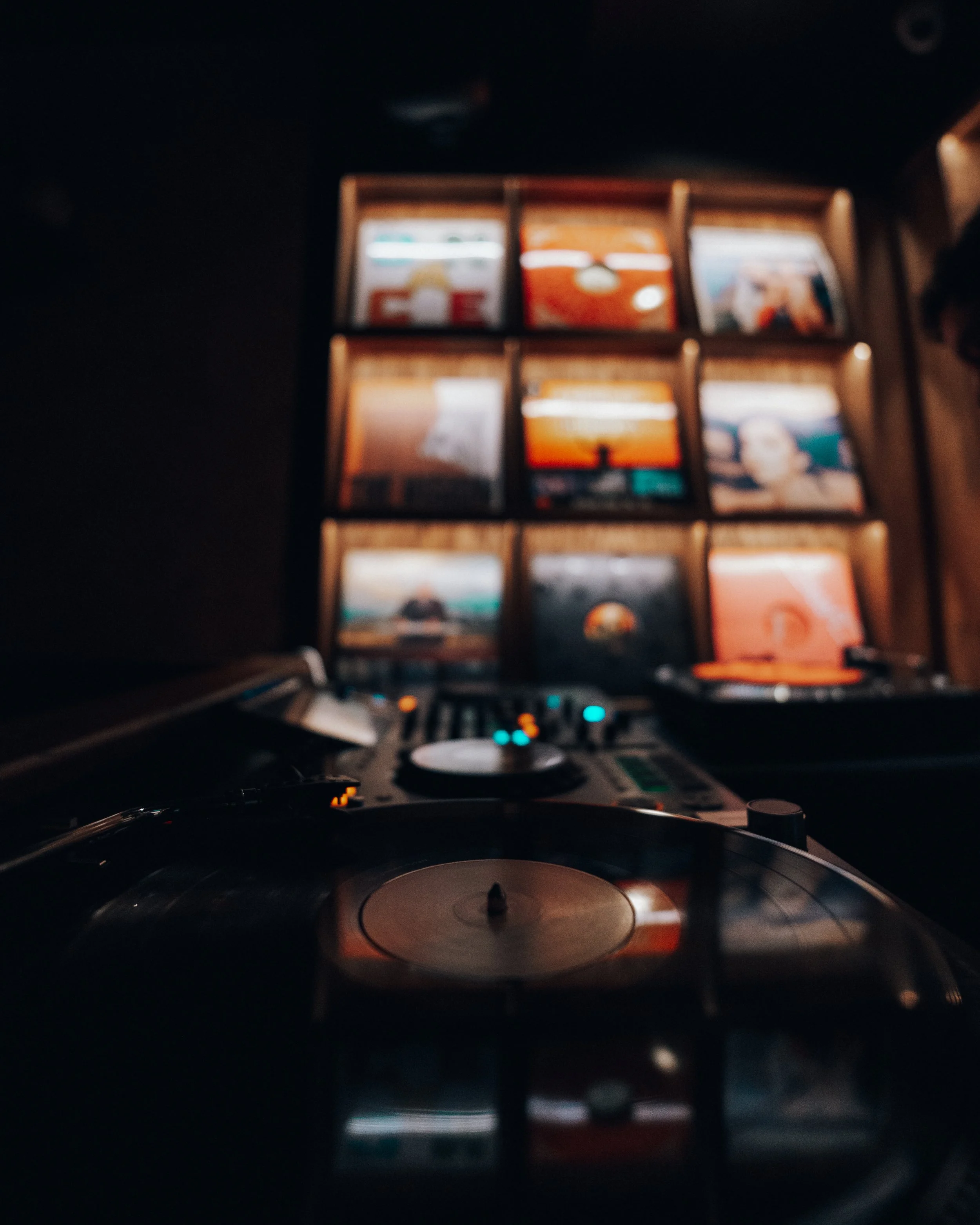 DJ turntable in a dark room with a collection of vinyl records displayed on a wall behind it.
