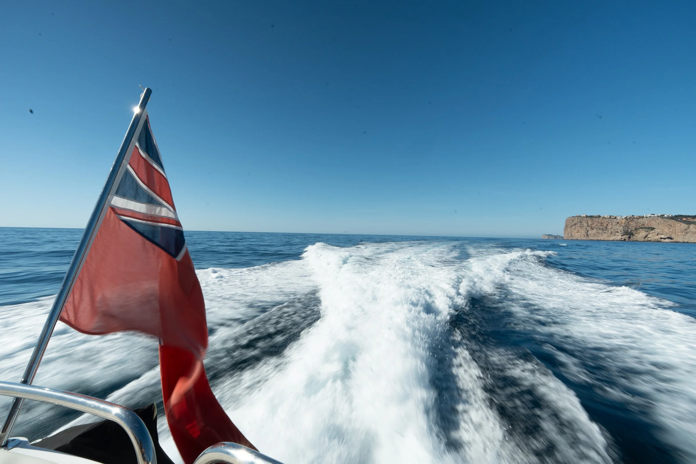 Photo of a boat moving on the ocean with a UK flag attached, a clear blue sky, and land visible in the distance.