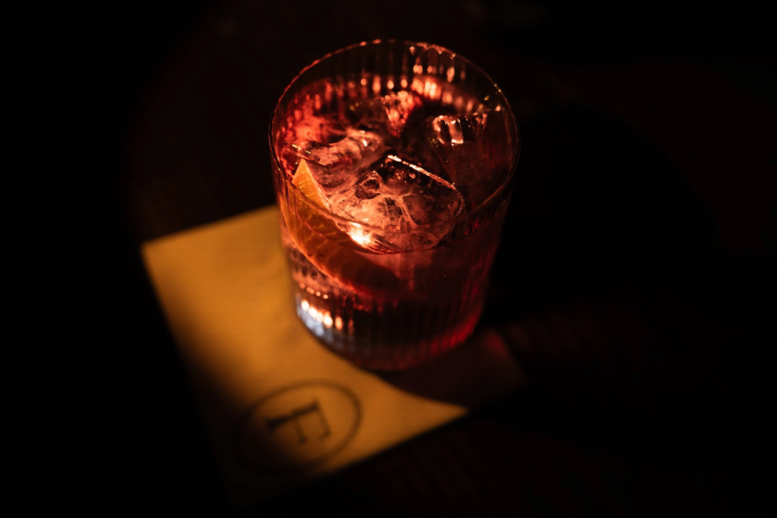 A glass of a pinkish-red beverage with ice cubes, sitting on a yellow piece of paper on a dark surface.