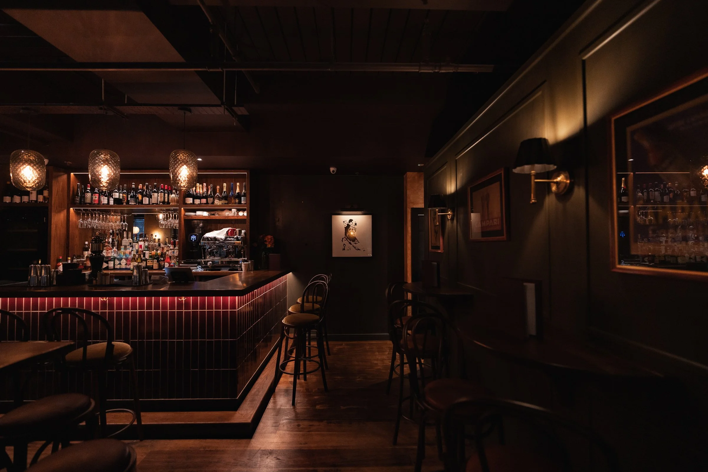 Dimly lit bar with a wooden countertop, bar stools, framed pictures on dark walls, and pendant lights hanging above the bar.